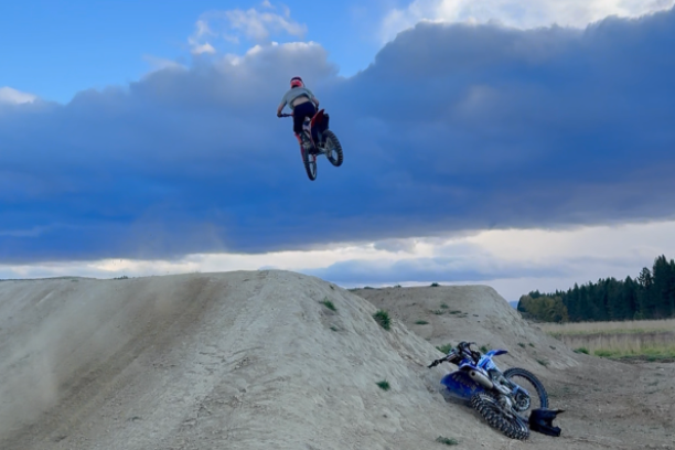 A person performing a jump on a mountain bike over a dirt jump, with a fallen mountain bike on the ground below, under a cloudy sky.