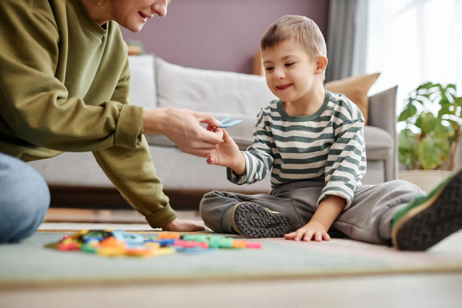 A young boy sitting cross-legged on the carpet while receiving a small object from an adult. The adult, partially visible, is smiling and wearing a green sweatshirt. Colored tokens or game pieces are scattered on the floor.