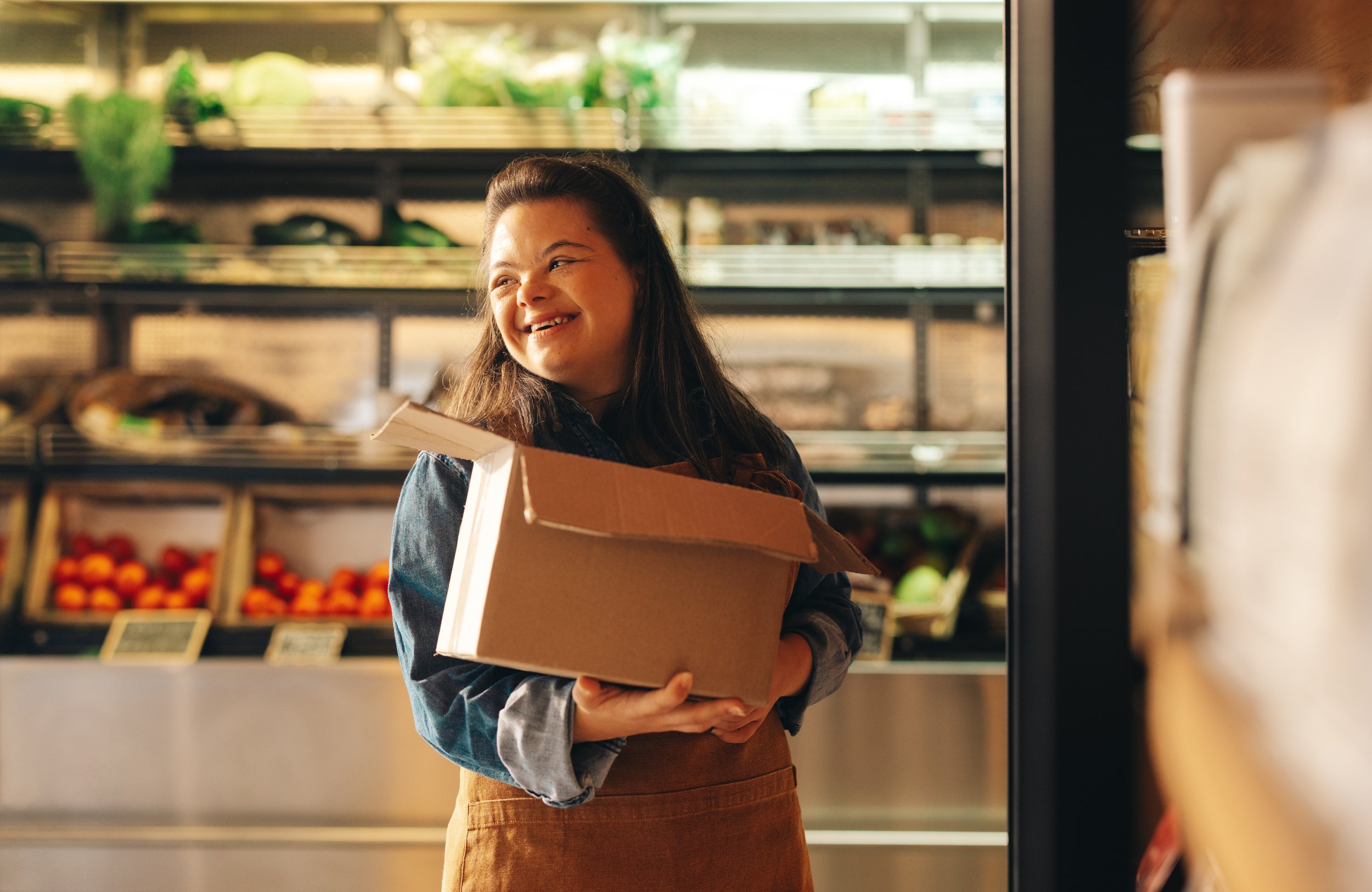 A young girl with long dark hair smiling while shopping in a grocery store, holding a cardboard box. Shelves of fresh produce are visible in the background.