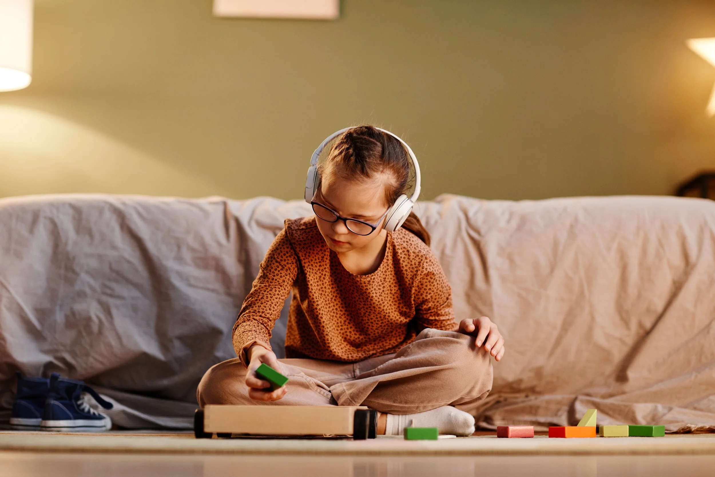A young girl with glasses and headphones sitting on the floor playing with colorful building blocks in a cozy living room.