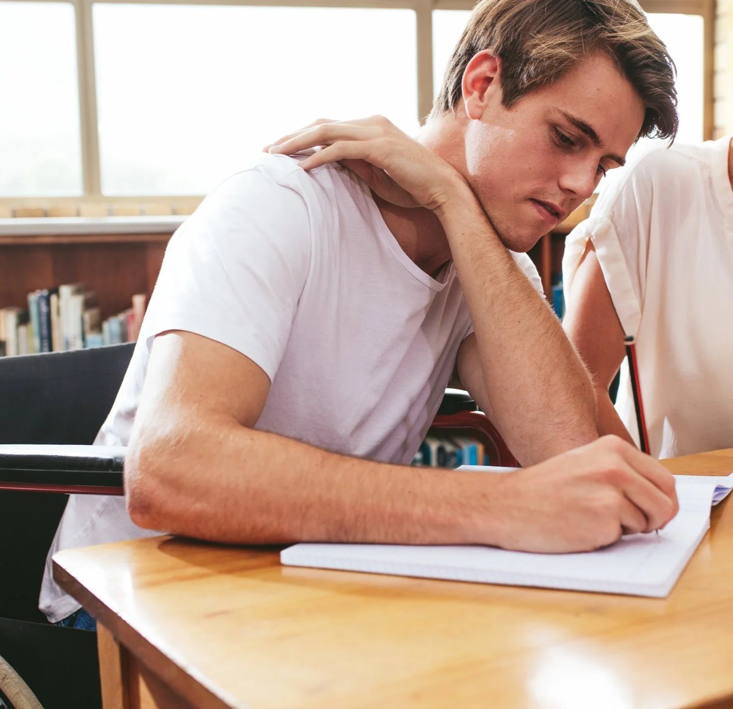 Young man in a white t-shirt writing in a notebook at a wooden table in a library.