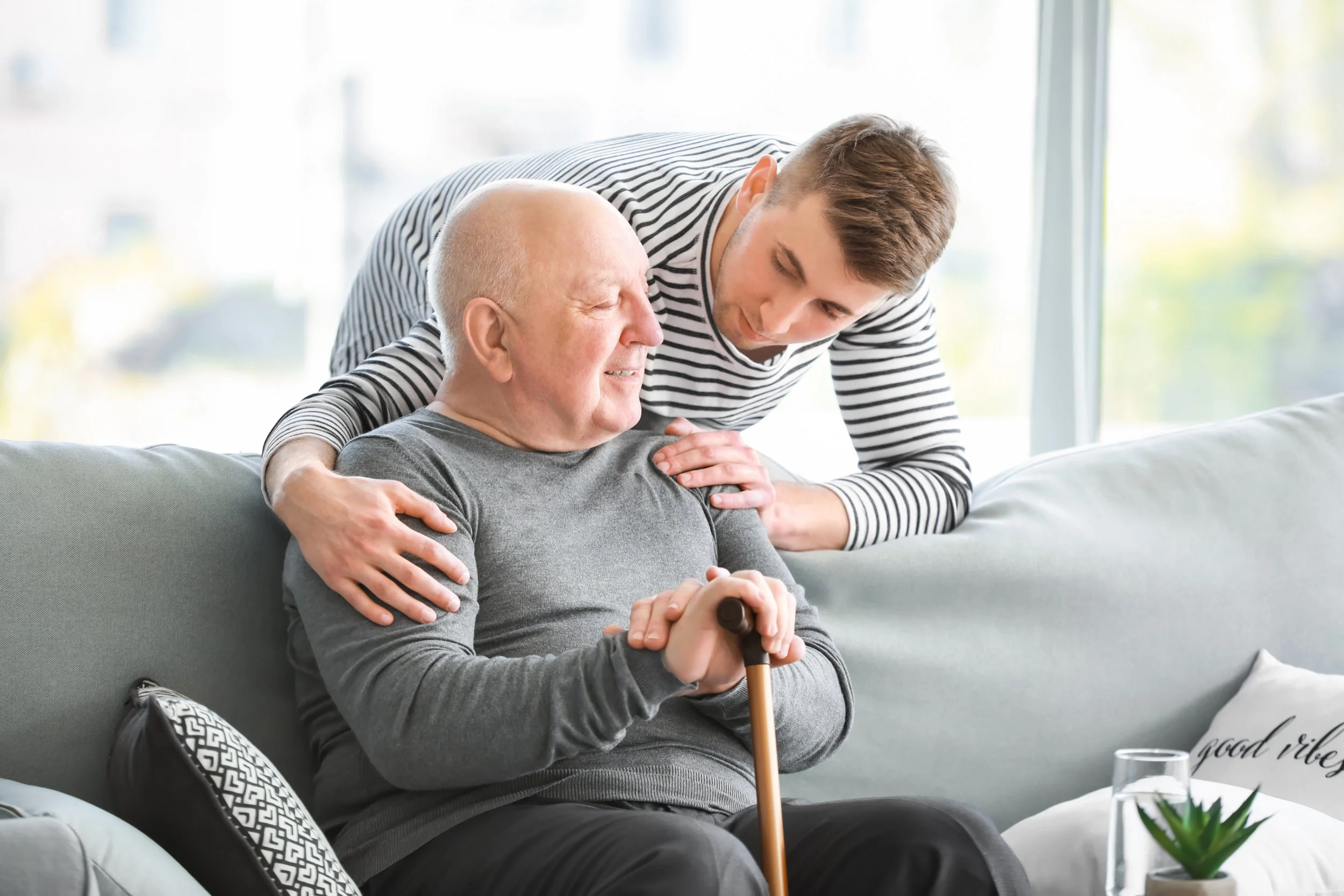 A young man and an elderly man engage in a caring moment in a bright living room. The elderly man, holding a cane, smiles, while the young man gently leans over and places a hand on his shoulder, showing support and compassion.