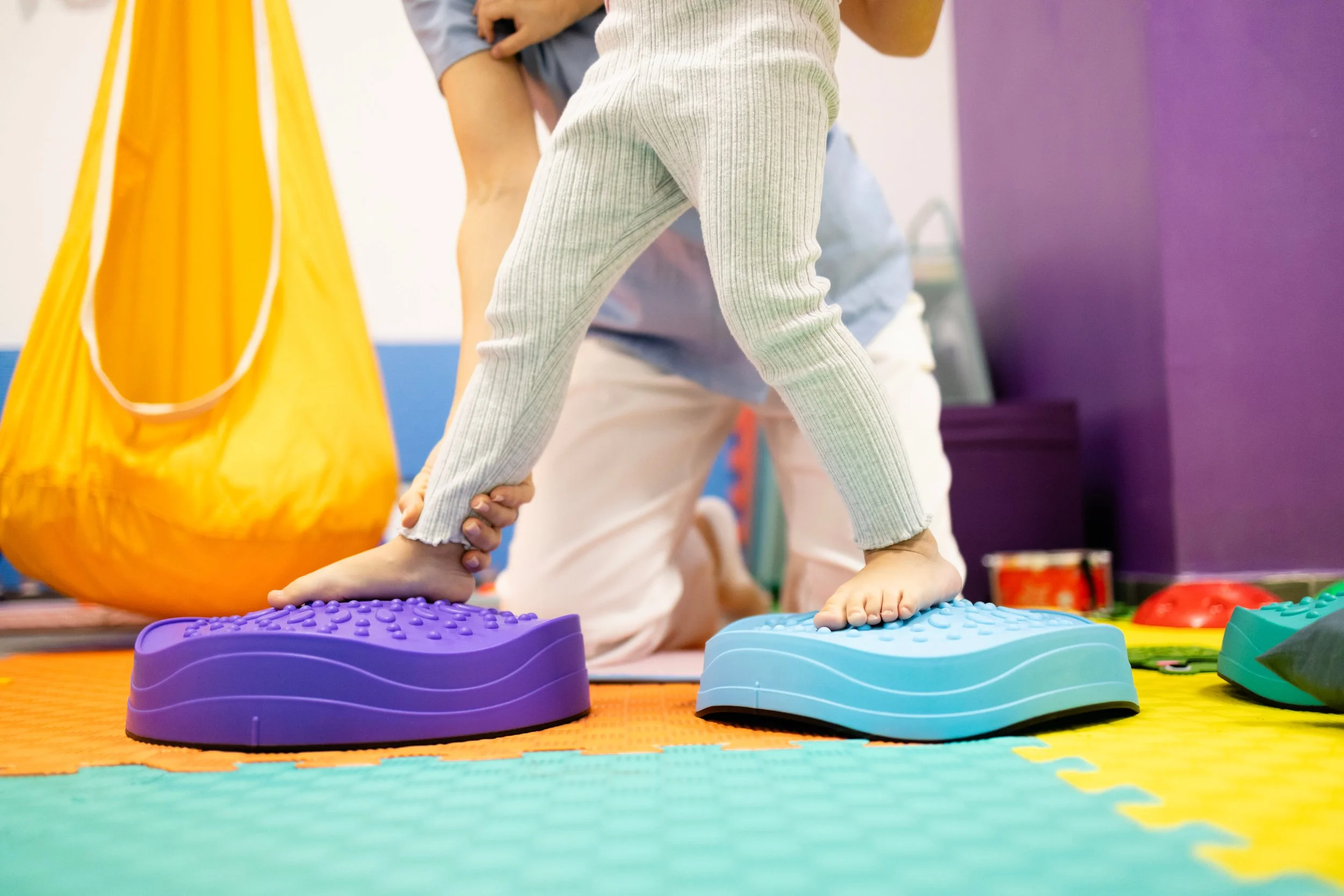 A child balancing on two colorful wobbly stepping stones, one purple and one blue, in a playroom with foam mats on the floor.