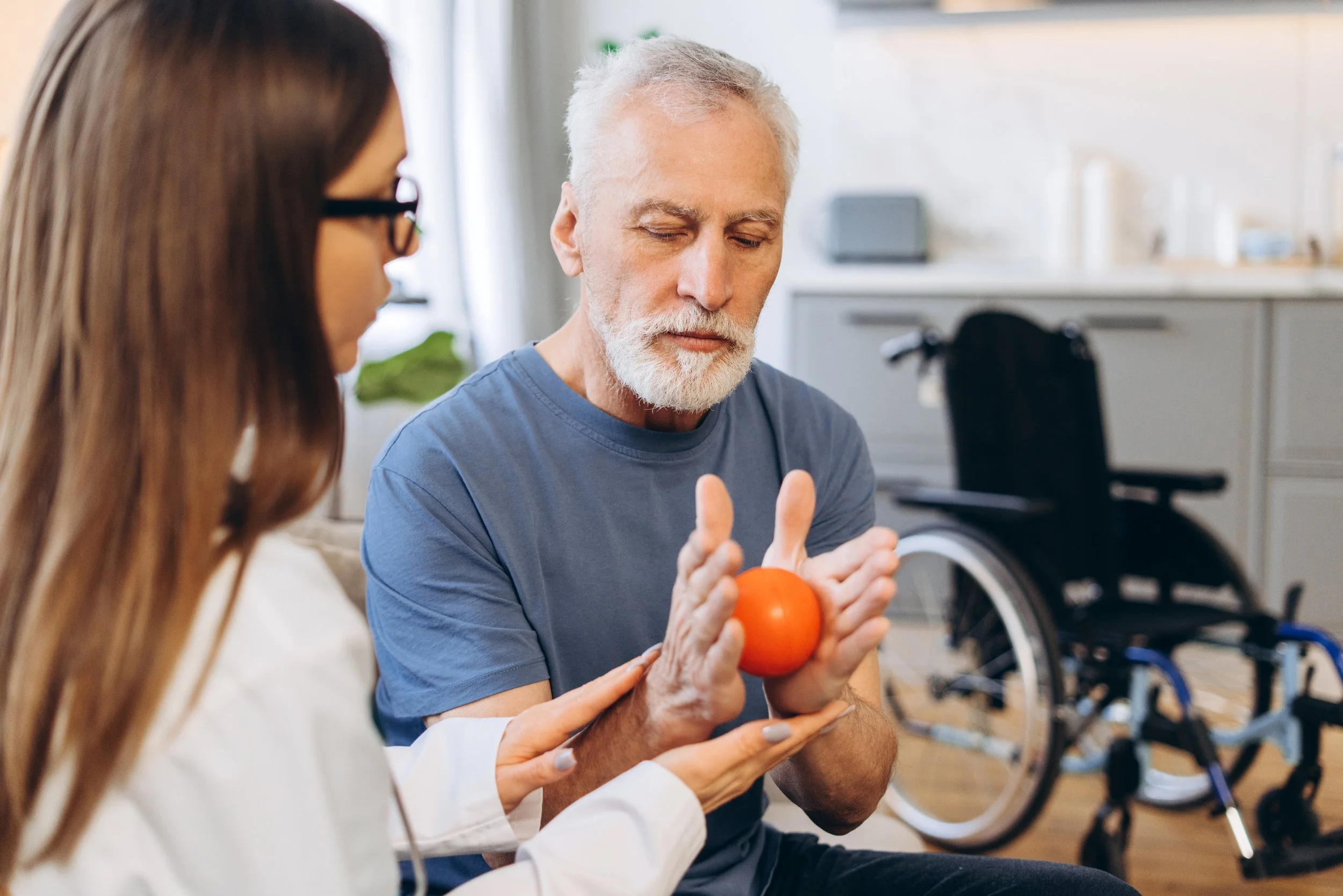 A man with a gray beard in a blue shirt sitting in a wheelchair receiving physical therapy with a therapist holding a red ball.