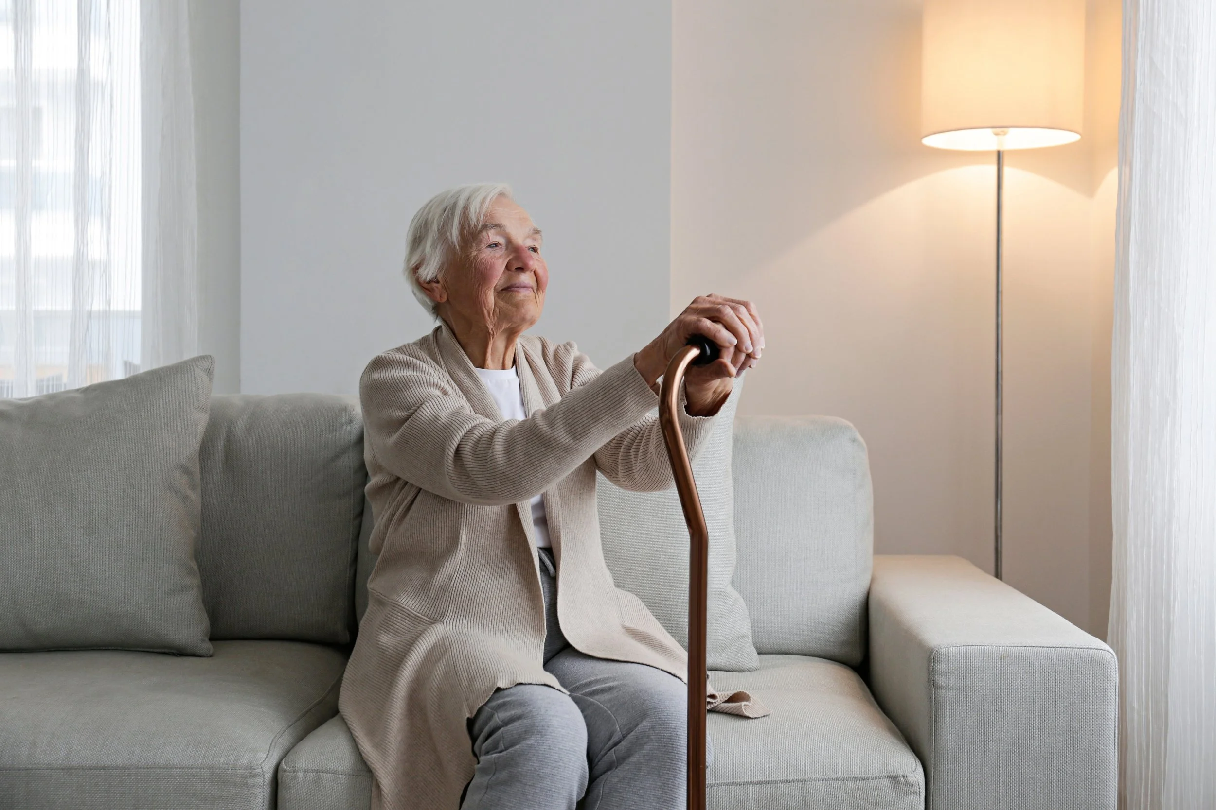 An elderly woman sitting on a light gray sofa, holding a wooden cane, with a content expression, next to a floor lamp and a sheer curtain in a well-lit room.