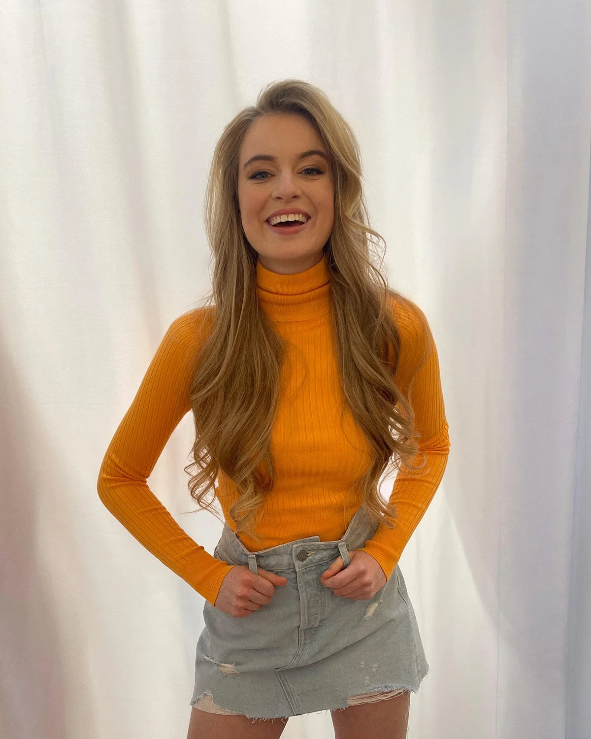 A young woman with long wavy hair, wearing a bright orange turtleneck and a distressed denim skirt, standing against a plain white backdrop with her hands on her hips and smiling.