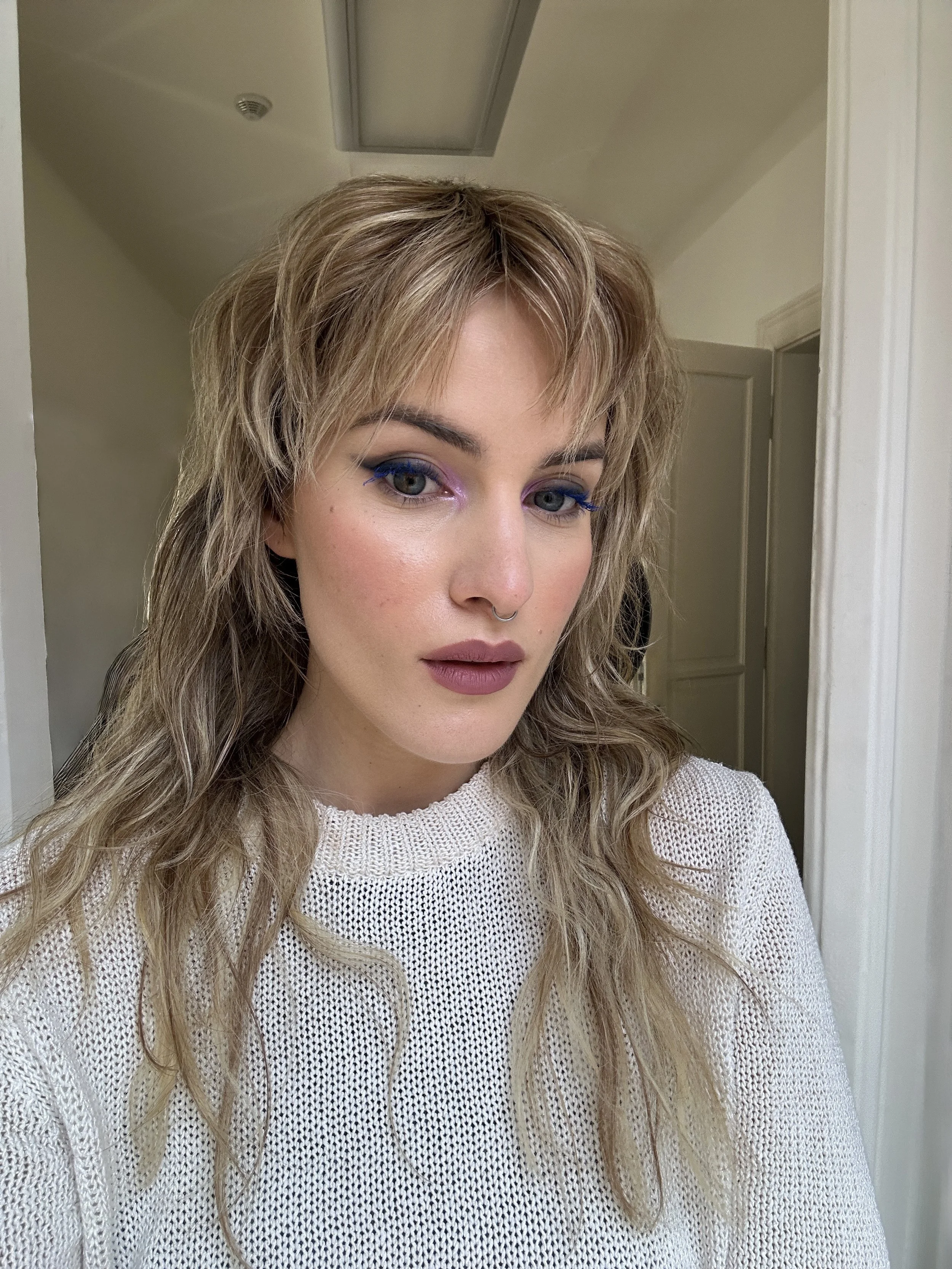 A woman with blonde, curly hair and makeup, wearing a beige knitted sweater, standing indoors near a doorway.