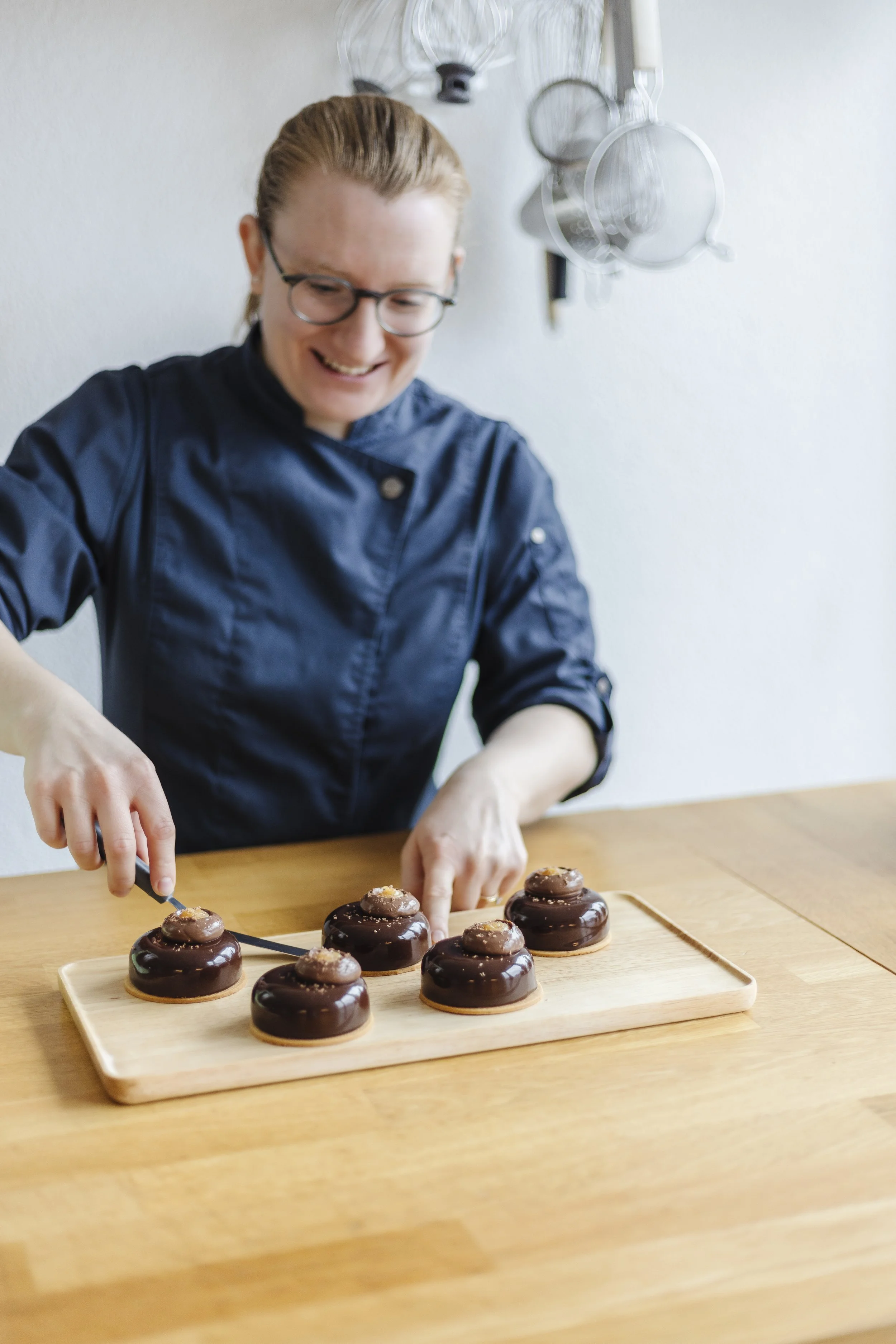 A person with glasses in a navy chef's coat is decorating chocolate desserts on a wooden tray, standing at a wooden table. There are kitchen utensils hanging on the wall in the background.