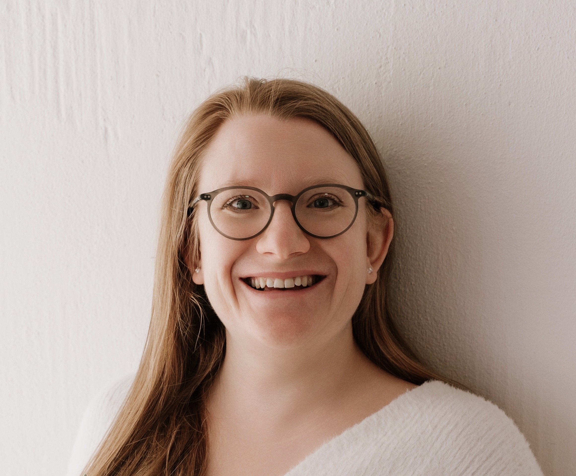 Close-up of a smiling woman with long red hair, glasses, and pierced ears, standing against a plain light-colored wall.