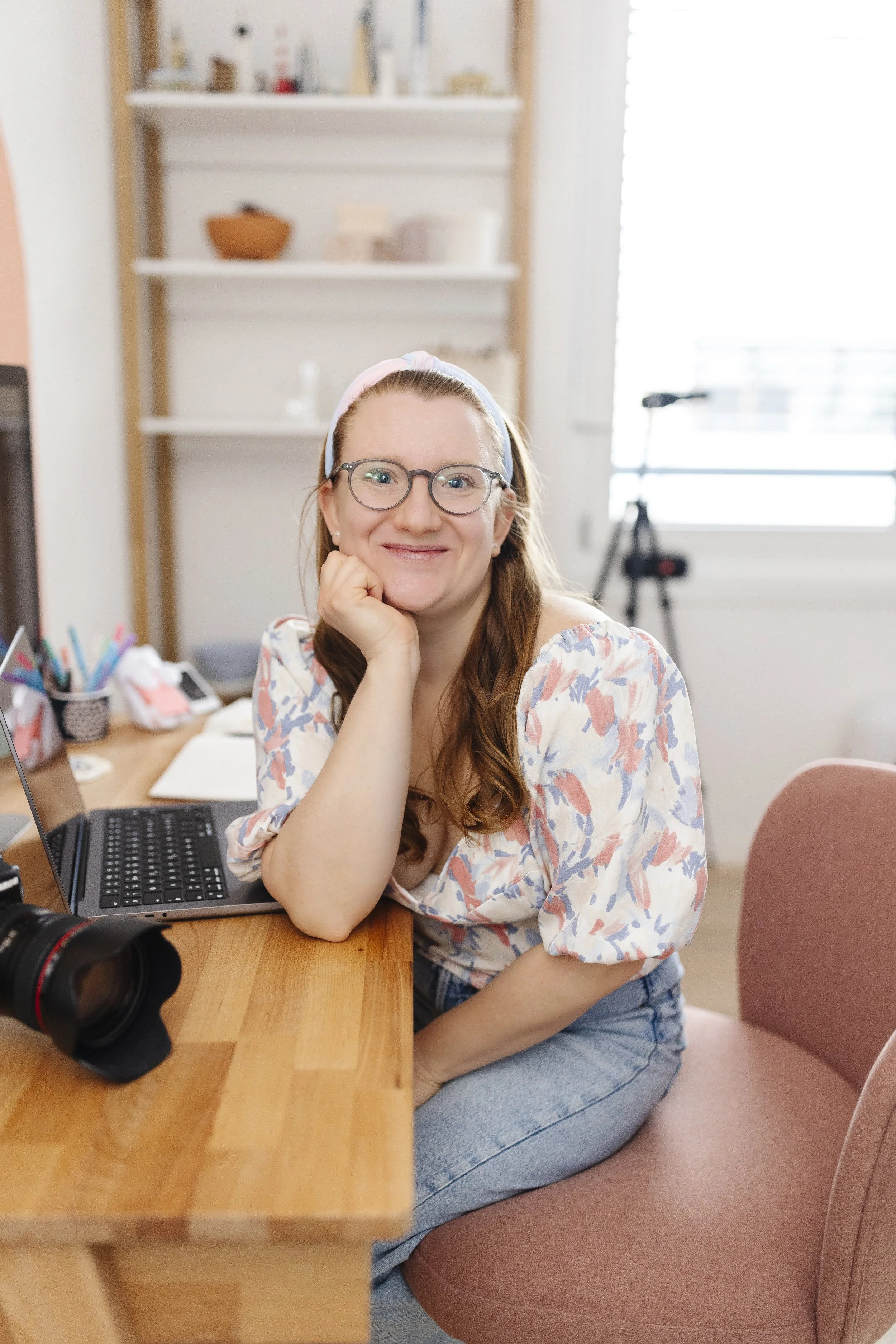 A woman with glasses and a headband smiling and resting her chin on her hand, sitting at a wooden desk with a laptop, in a bright room with shelves and a window in the background.