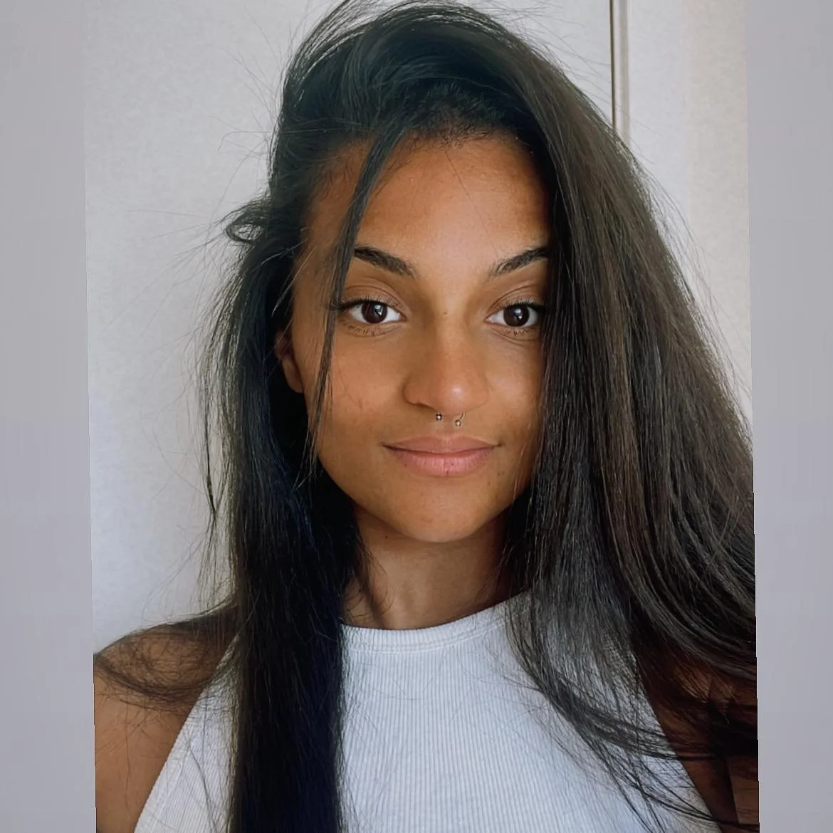 Jeune femme avec des cheveux longs et noirs, portant un t-shirt blanc, regardant l'objectif avec un léger sourire, fond neutre.