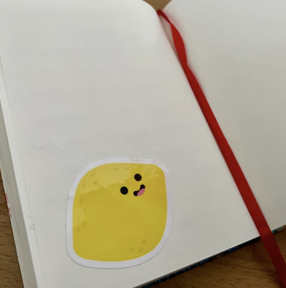 Offene weiße Buchseite mit einem gelben Sticker in Form eines quadratischen Charakters mit einem lachenden Gesicht, und eine rote Stoffmarkierung an der Seite.