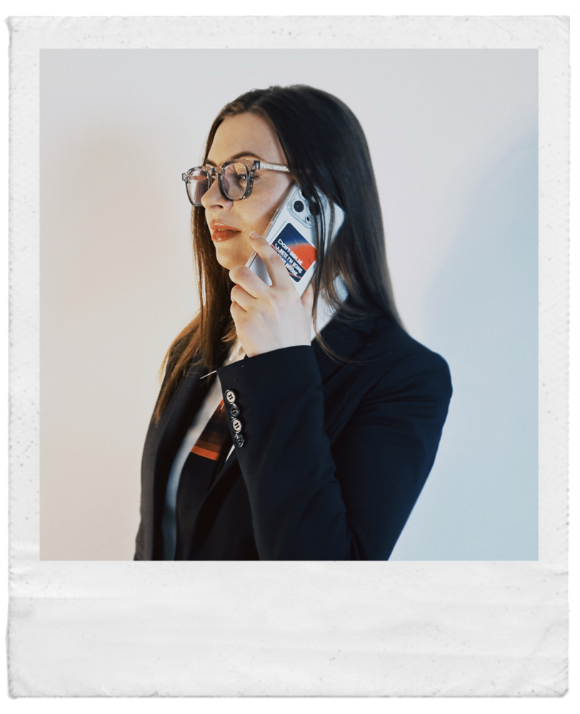 Frau in schwarzer Kleidung mit Brille und langen braunen Haaren spricht am Smartphone.