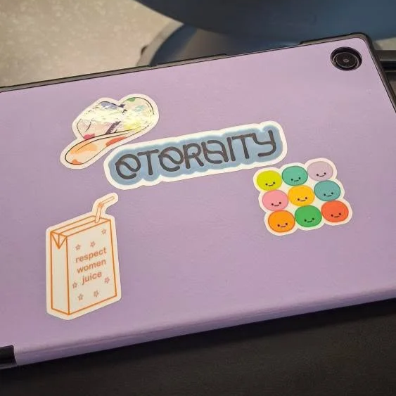 Laptop mit Sticker, der 'OTORACITY' schreibt, bunte Smileys, eine Tüte mit der Aufschrift. 'respect women juice' und eine kleine Illustration eines Huts.