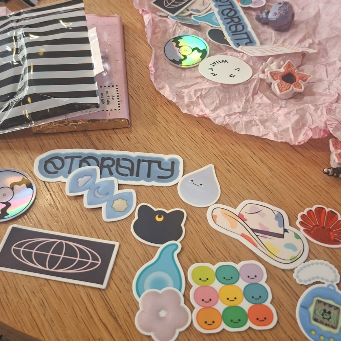Verschiedene bunte Sticker und kleine Plüschfiguren liegen auf einem Holztablett, darunter eine lachende Wolke, bunte Kugeln, ein Loewe-Symbol, ein Tropfen, ein liegender Typ mit buntem Muster, und andere dekorative Aufkleber.