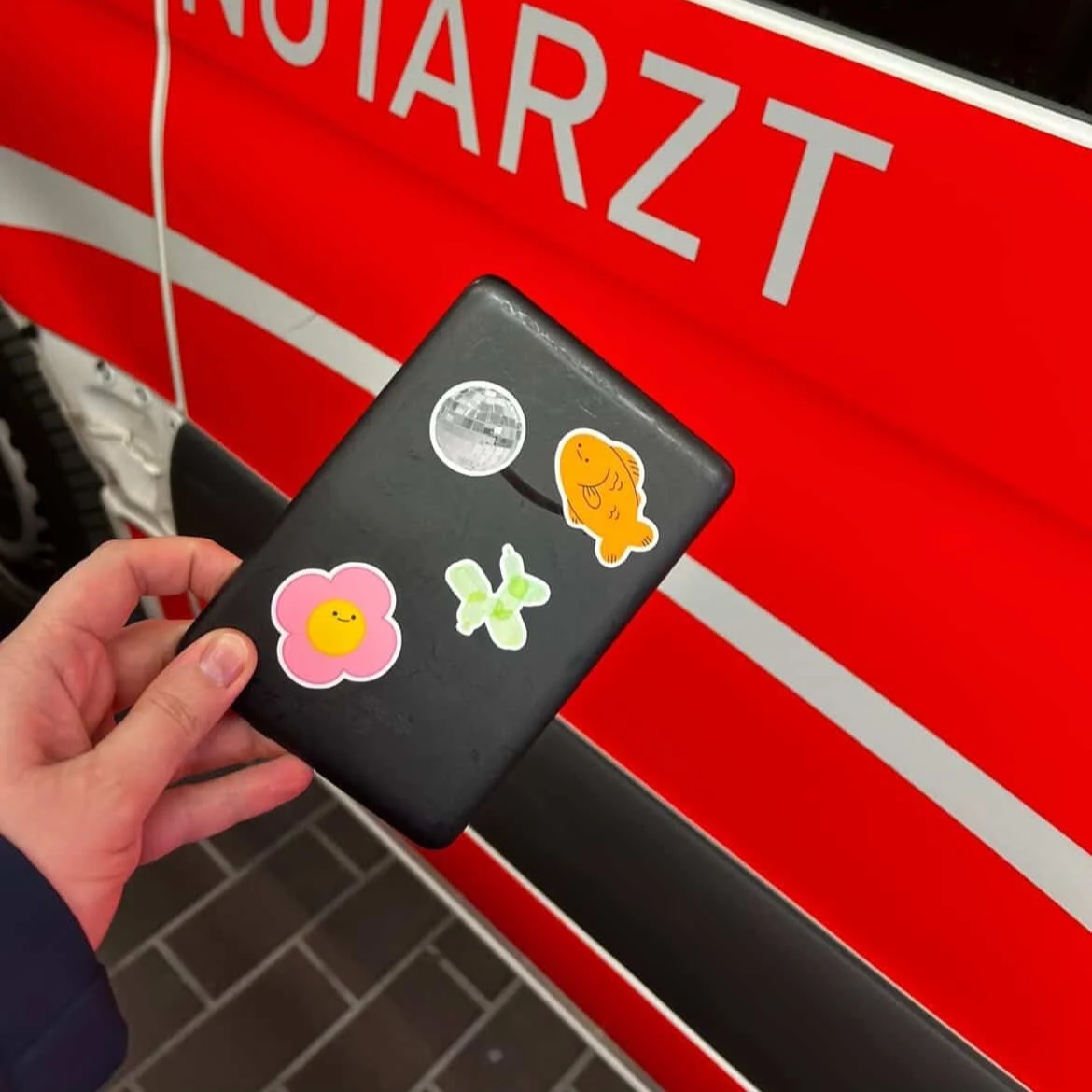 Eine Hand hält eine schwarze Brieftasche mit vier bunten Sticker vor einem roten Feuerwehrfahrzeug, auf dem das Wort 'NOTARZT' zu lesen ist.