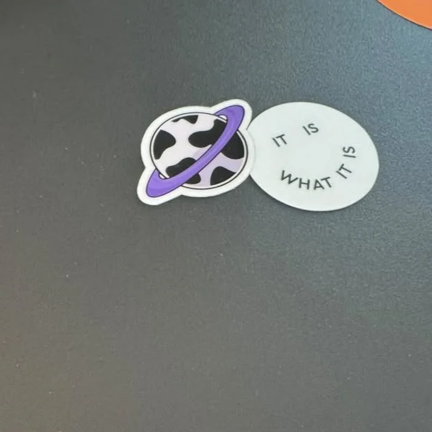 Auf dem Bild sind zwei Aufkleber, einer mit einem Planeten und einem Ring um ihn, der andere mit dem Text "IT IS WHAT IT IS" auf einem dunklen Hintergrund.