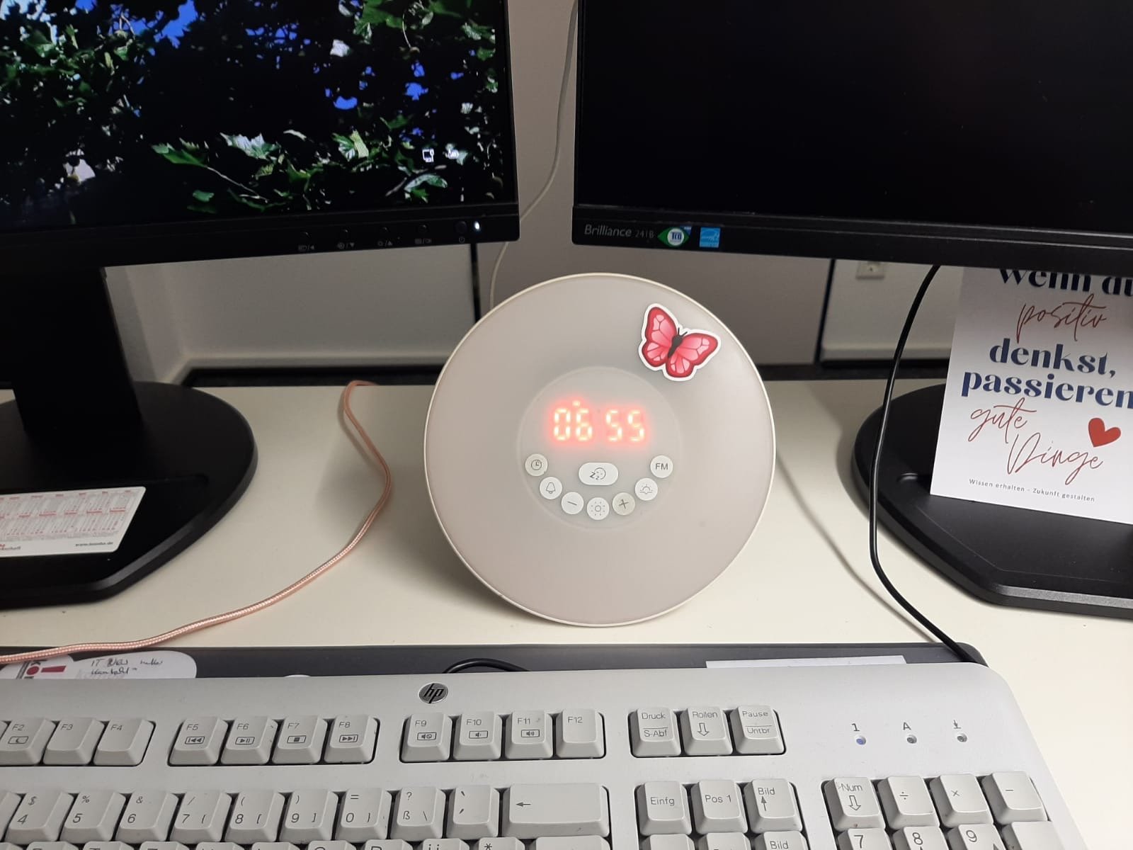 Ein Schreibtisch mit zwei Computerbildschirmen, einer Uhr mit Beleuchtung und einem Herz-Butterfly-Sticker, daneben ein Poster mit deutschen Sprüchen und einem Herz. Außerdem eine weiße Tastatur im Vordergrund.