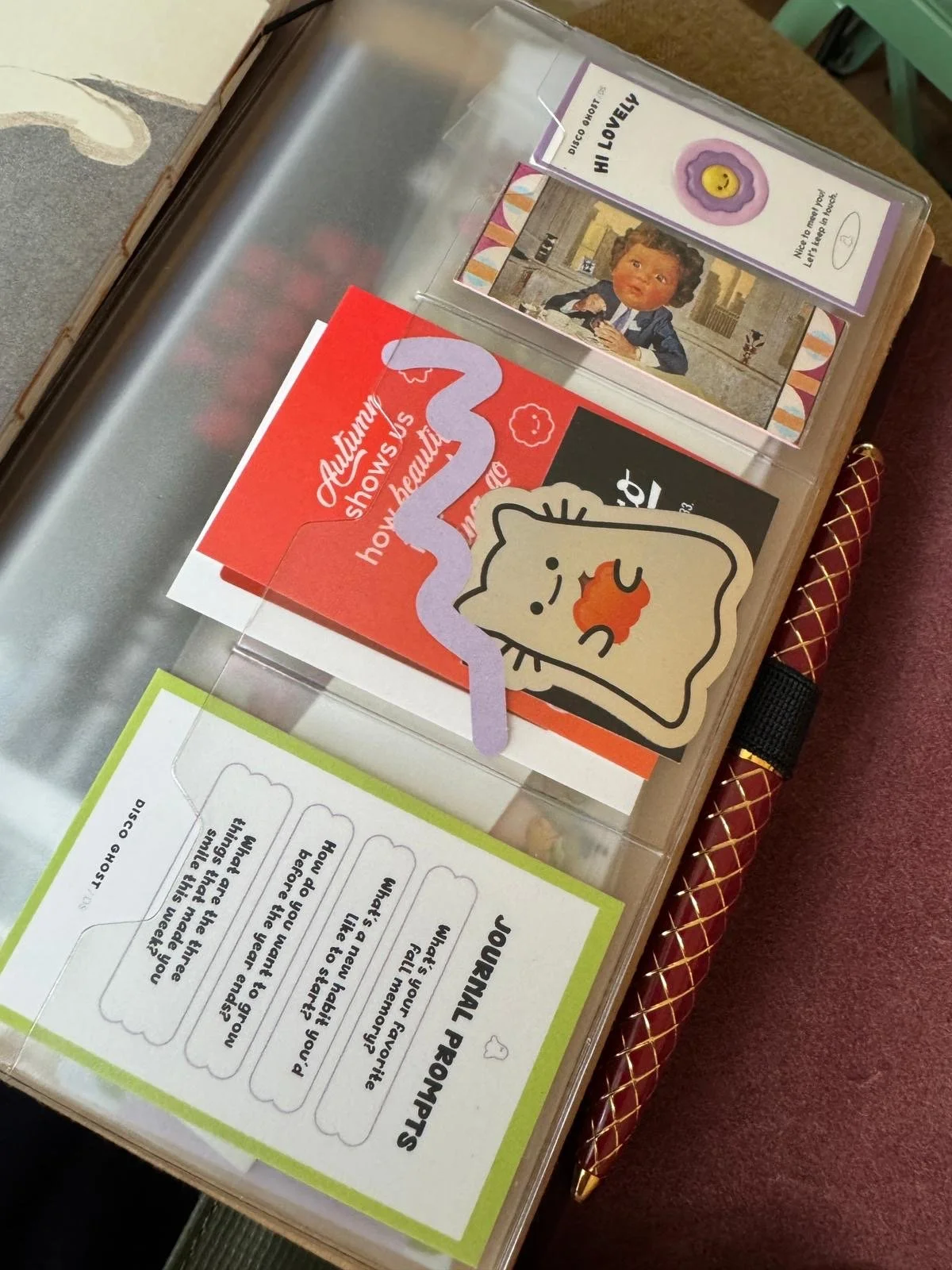 Ein Tisch mit verschiedenen Kärtchen und Notizzetteln, darunter eine Karte mit einem lachenden Stickers mit dem Text 'HI LOVELY' und einer Illustration einer Person, sowie eine orange Karte mit dem Text 'Autumn shows us how beautiful it can be to let