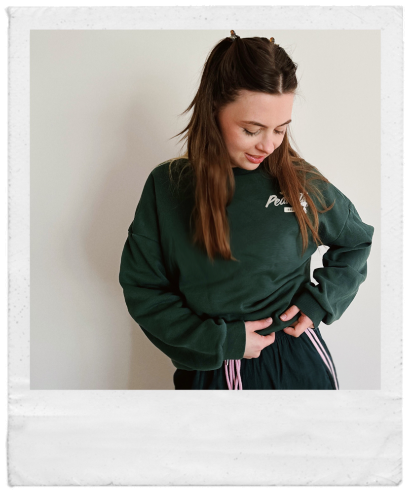 Ein junges Mädchen mit braunen Haaren trägt eine grüne Sweatshirt und eine grüne Hose mit pinkfarbenen Zughosen, schaut nach unten und hält den Bauch mit einer Hand, vor einem weißen Hintergrund.