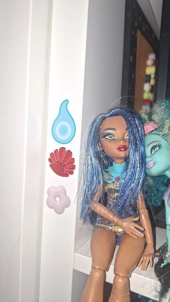 Dies ist ein Foto eines Puppenregals mit einer bekannten Monster High Puppe mit blauer und schwarzer Haarfrisur und einer weiteren, unvollständigen Puppe im Hintergrund.