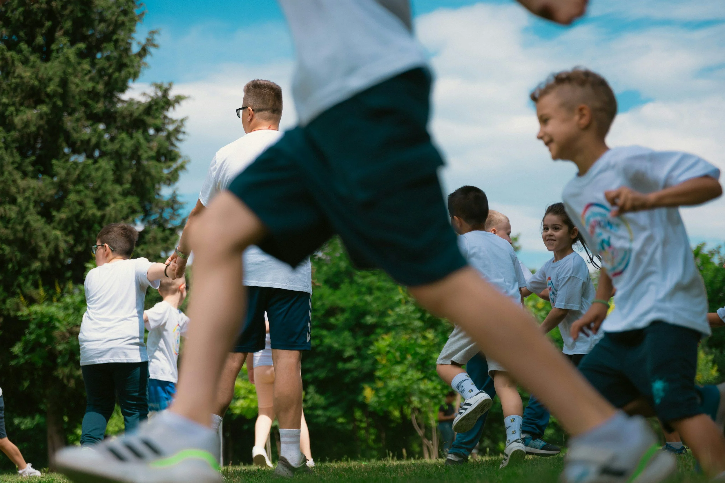 Kinder und Erwachsene, die im Freien an einem Gruppenspiel oder einer Aktivität teilnehmen, umgeben von Bäumen, bei schönem Wetter.