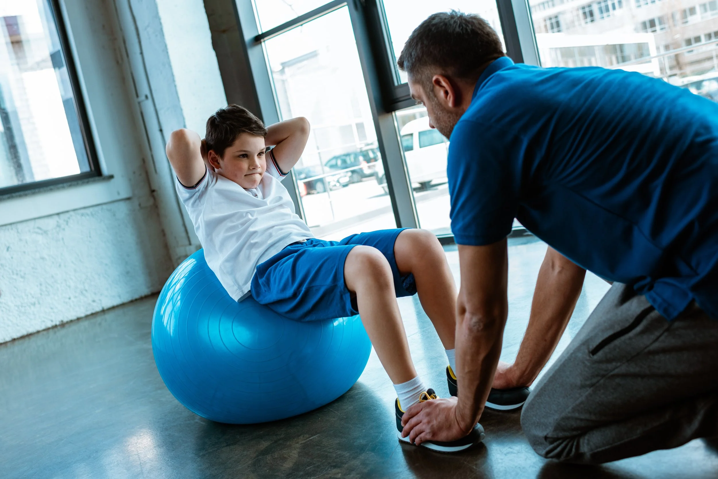 Ein Junge sitzt auf einem Fitness-Ball, während ein Trainer ihn beim Dehnen unterstützt in einem Fitnessstudio mit großen Fenstern.