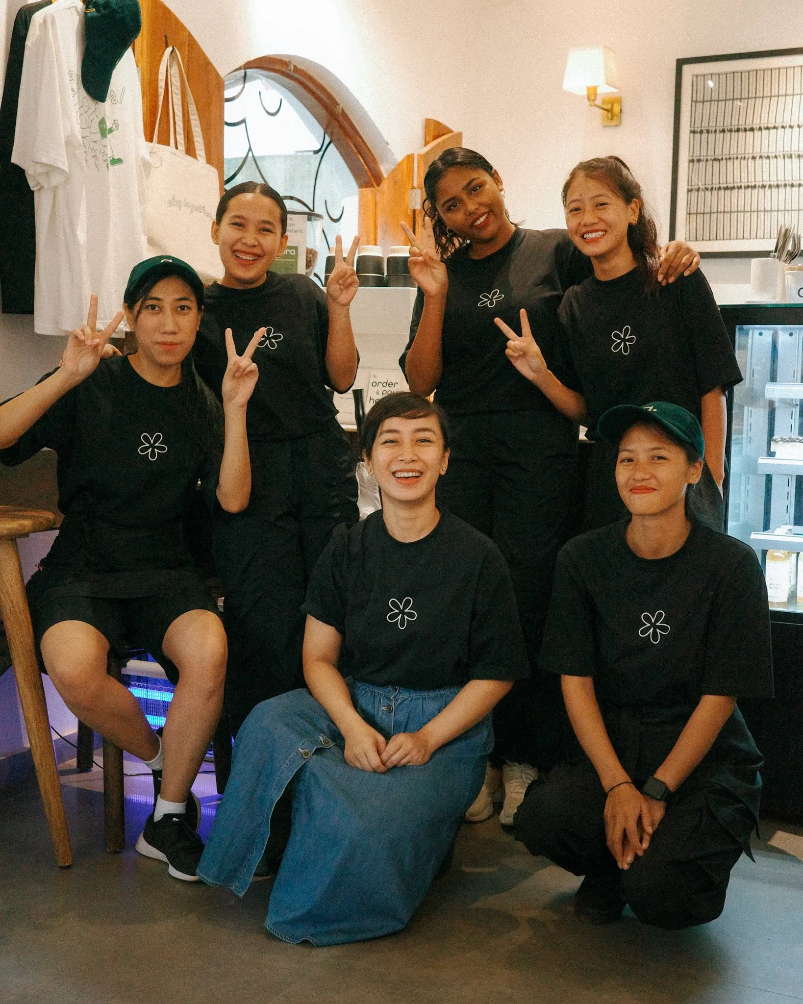 Six women in black shirts posing inside a restaurant, some making peace signs, smiling, with a window and food-related items in the background.