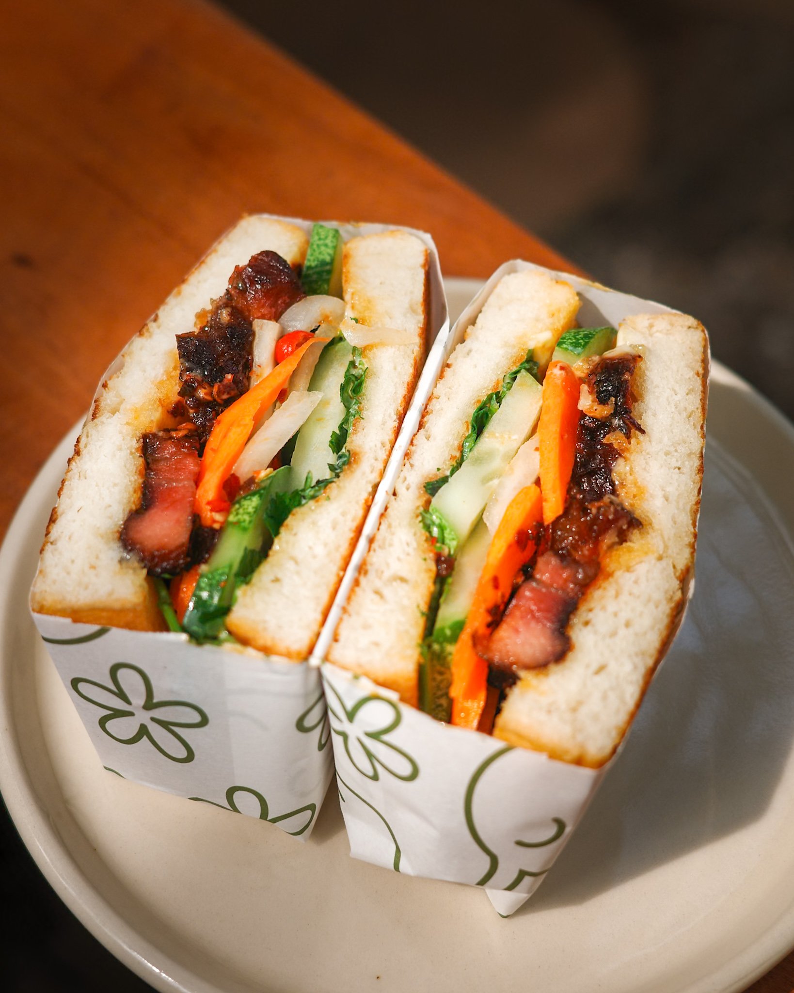 Two sandwich halves with bacon, lettuce, tomato, cucumber, and carrots in white bread, in decorative paper sleeves on a white plate.