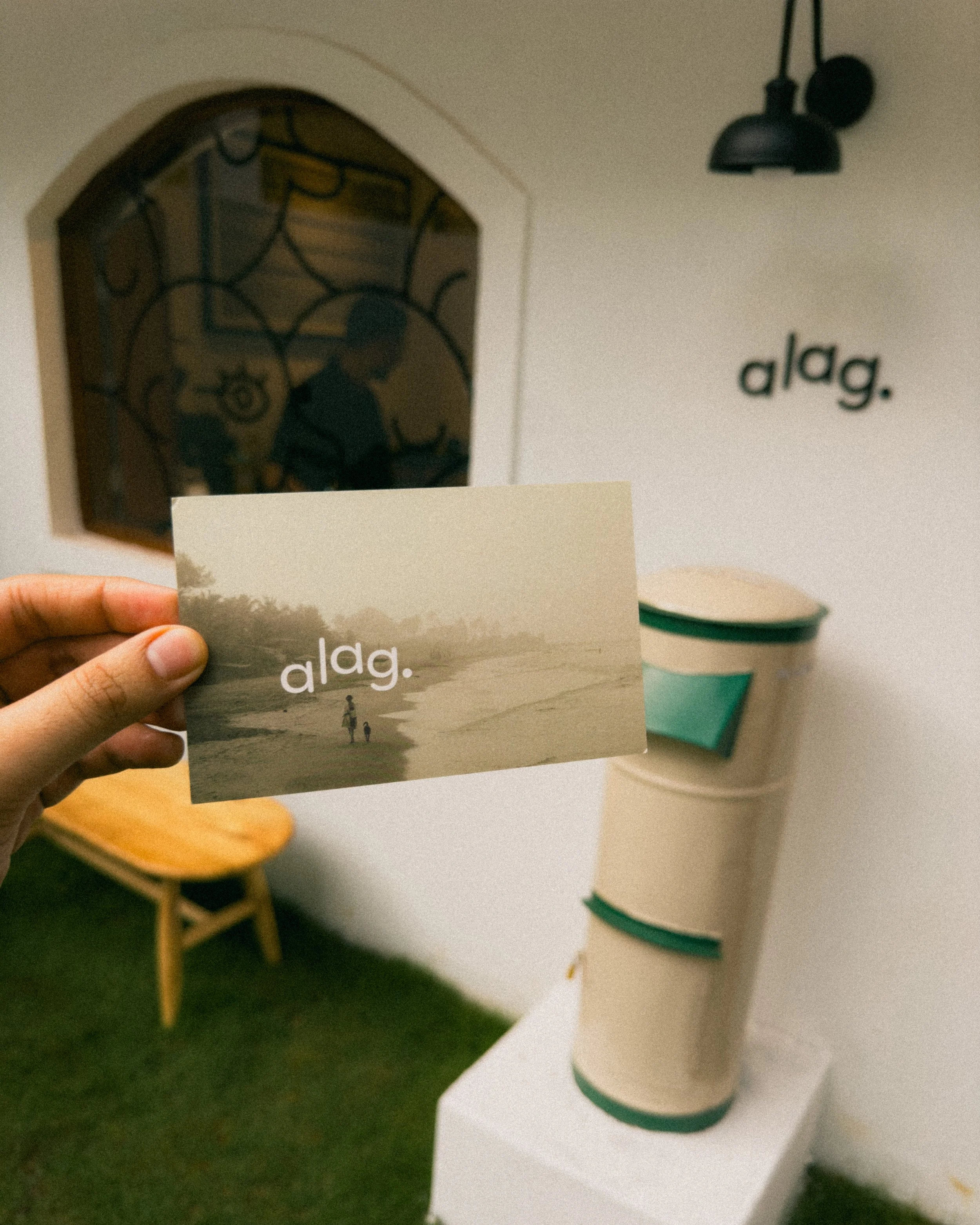 Person holding a card with the word 'a lag.' on it, in front of a display with the same word, 'a lag.', on a wall and a cardboard cutout of a beverage container.