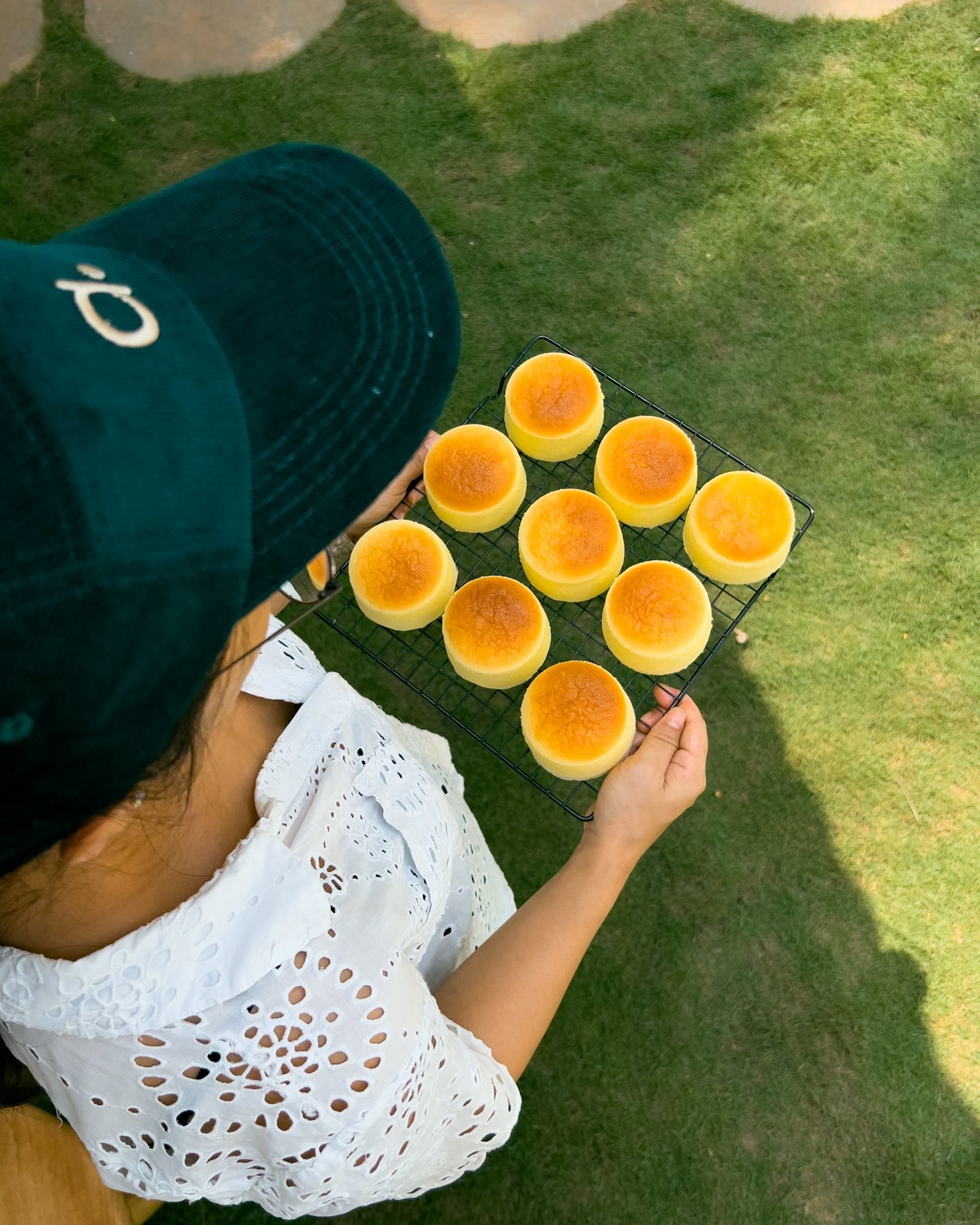 A person holding a wire rack with ten baked cheesecakes outdoors on green grass, wearing a white eyelet dress and a dark green cap.