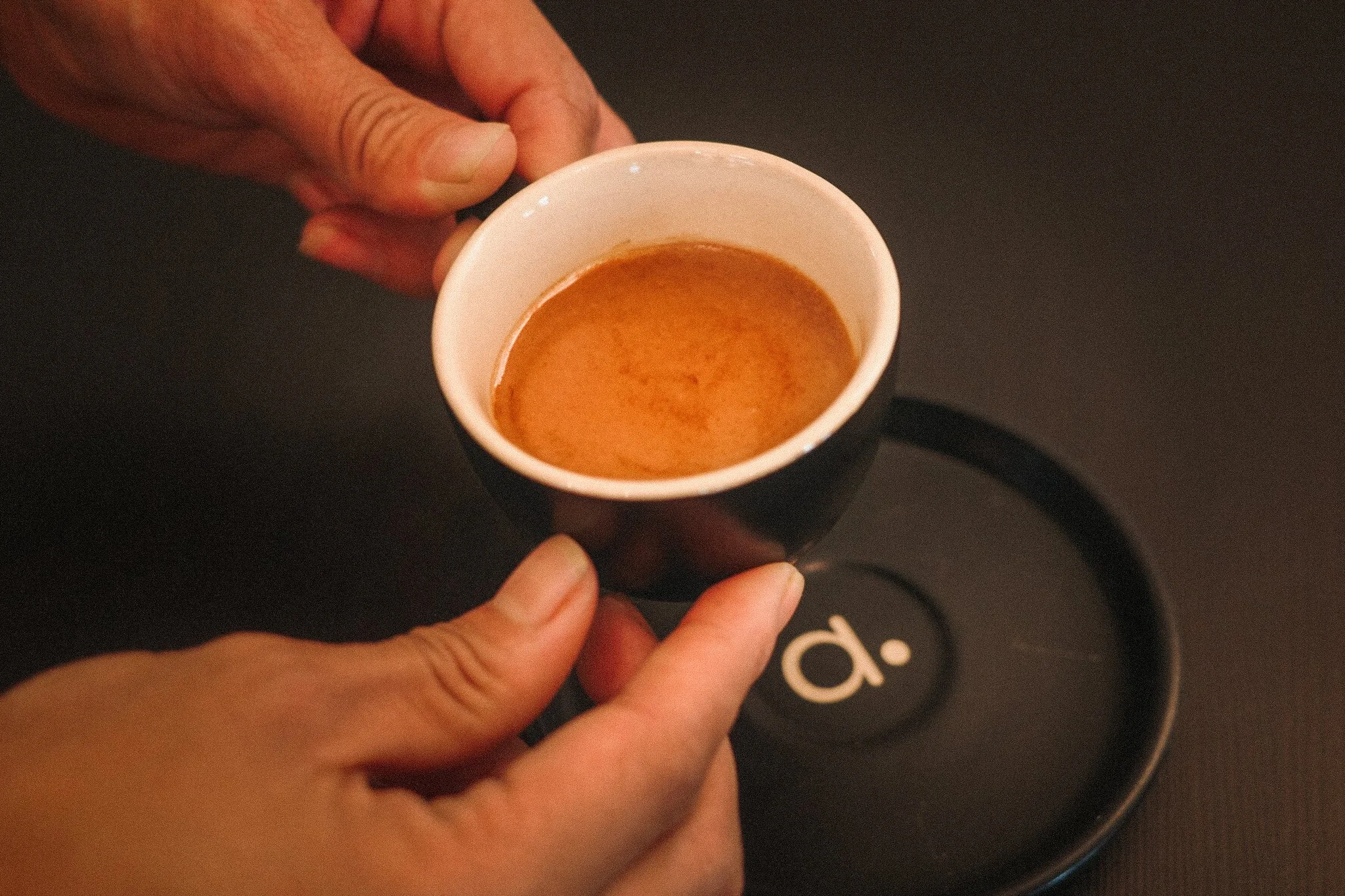 Person holding a cup of espresso with a black 'a.' on a coaster on dark surface.