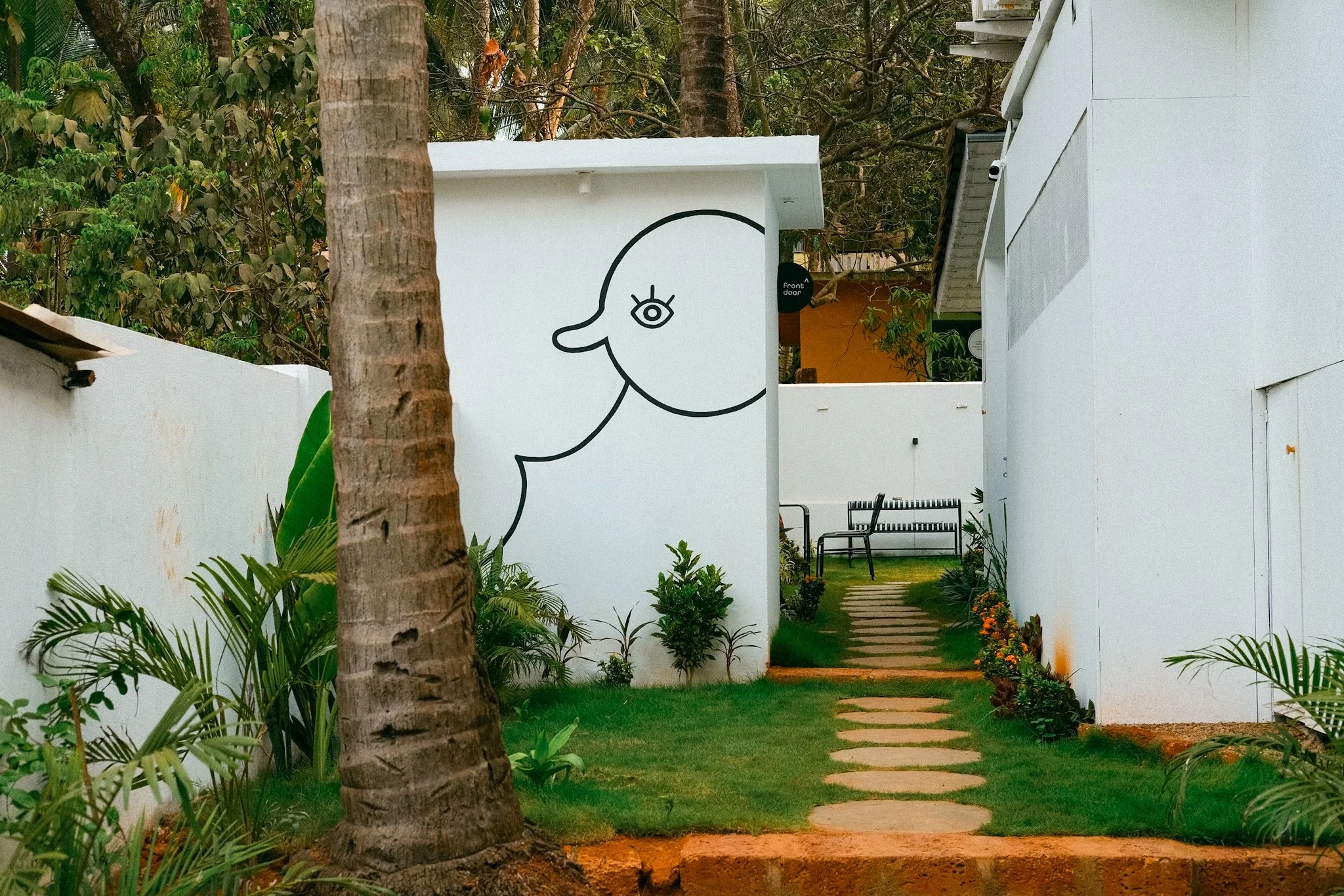 A pathway with stepping stones flanked by greenery and white walls, leading to a seating area in a garden. A large tree trunk is in the foreground, and a black line drawing of a stylized animal face is painted on the wall.
