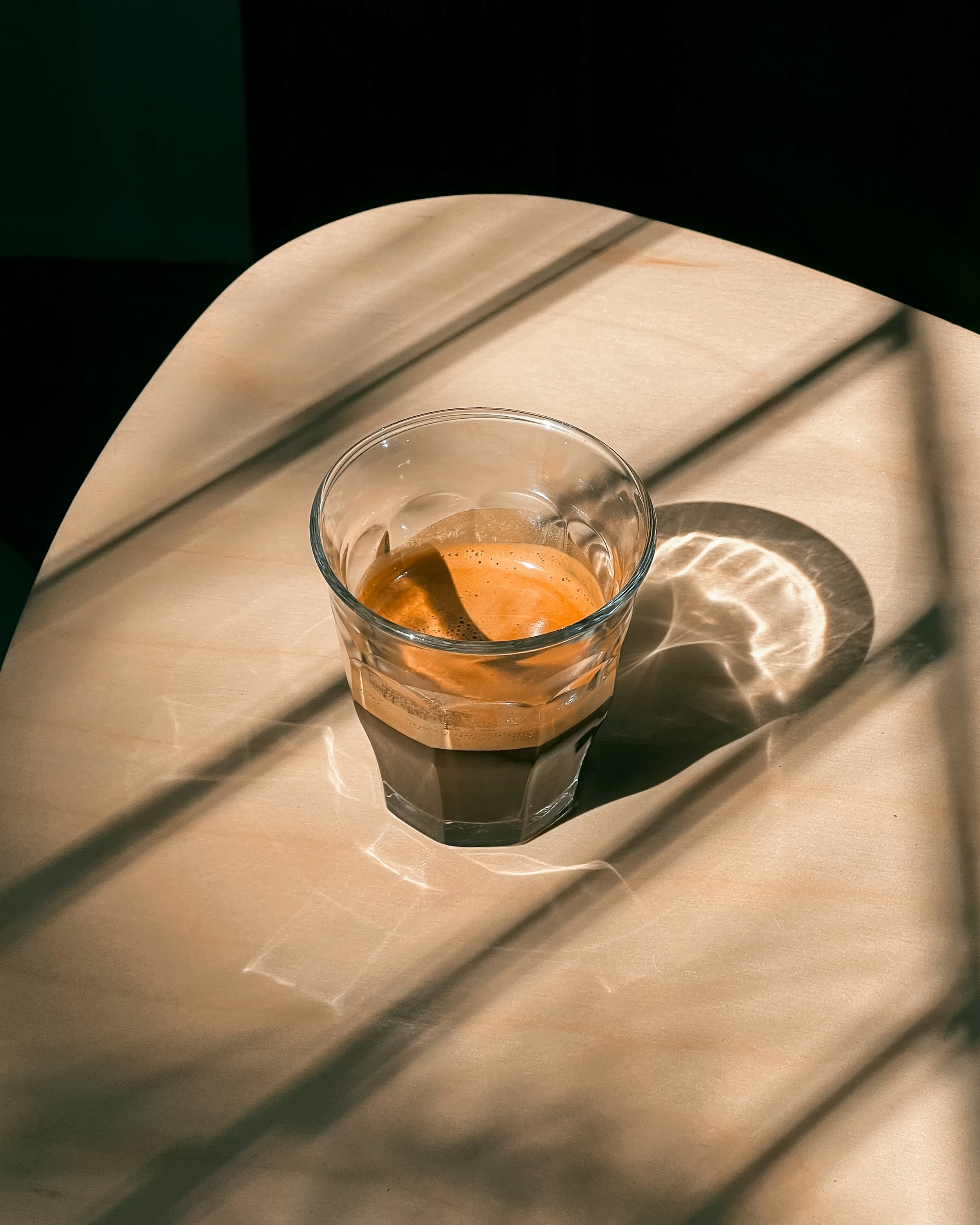 A glass of espresso on a light wood table with sunlight creating a reflection and shadow patterns.