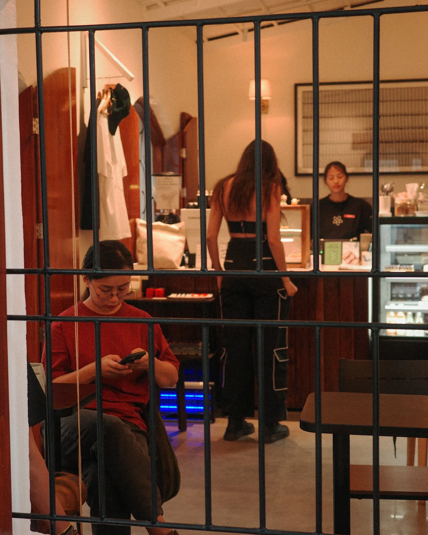 A view of a cafe or restaurant with three women, one seated looking at her phone and two working behind the counter, through a black metal grid.