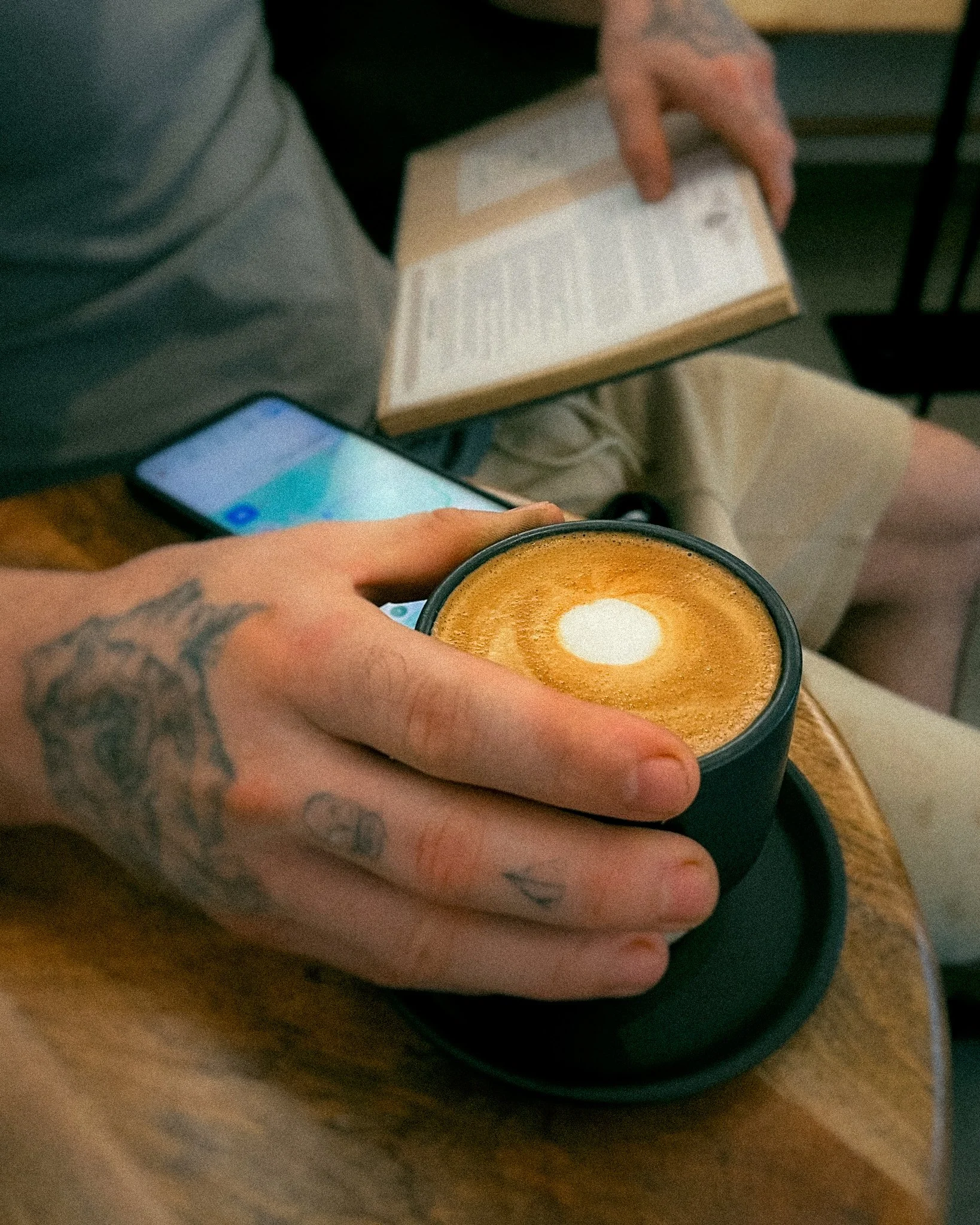 A person's hand with tattoos holding a black coffee cup with latte art on a wooden table.