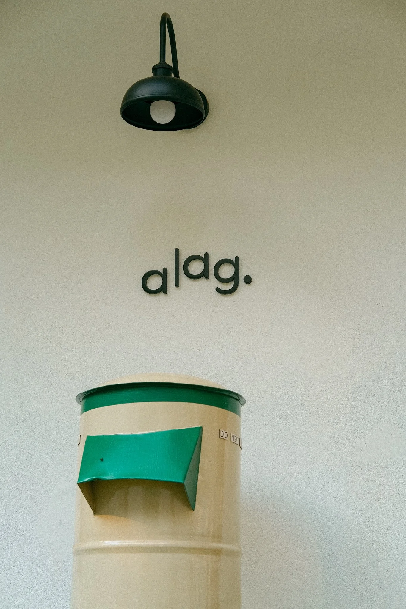 A wall-mounted lamp above a sign that reads 'a l g.' and a green mailbox with a torn flap.