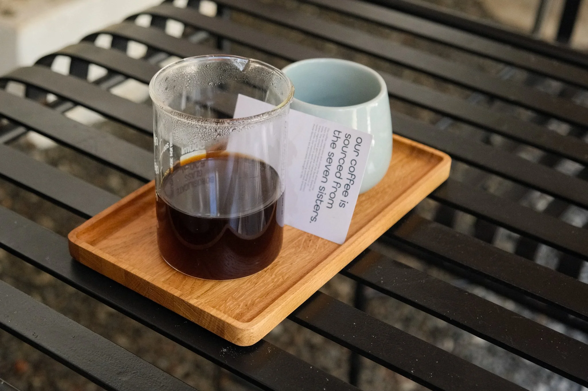 A wooden tray holding a glass of cold brew coffee, a light blue ceramic cup, and a printed note, all placed on a black metal outdoor table.