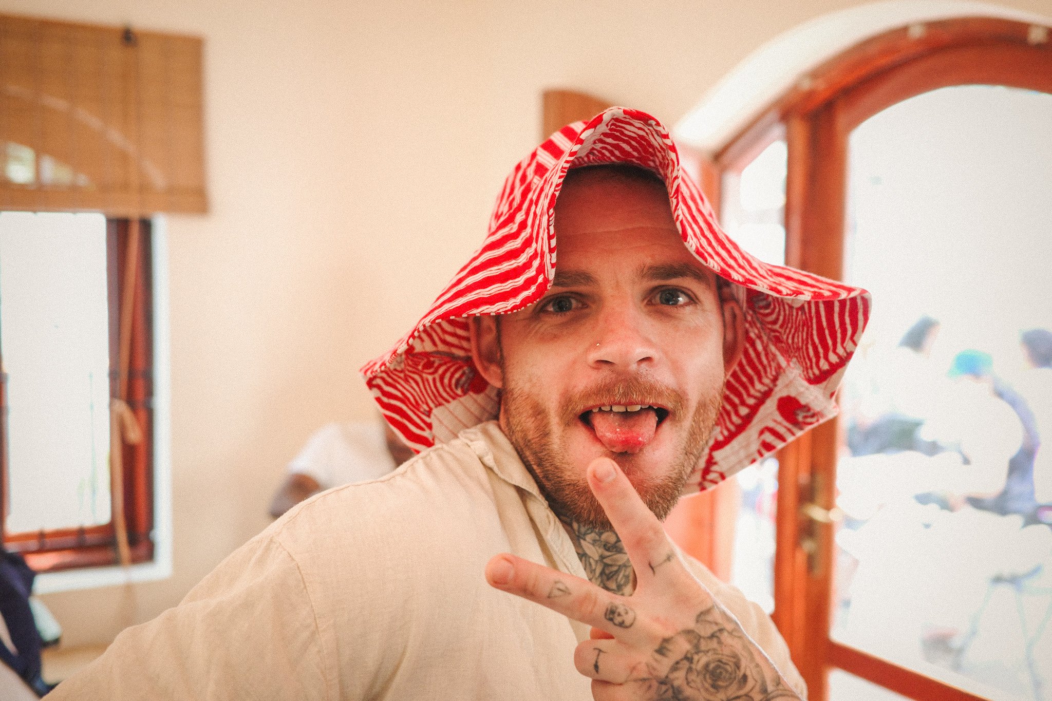 A man with a beard and tattoos making a peace sign with his hand, wearing a red and white striped sun hat, and sticking his tongue out inside a room with natural light.