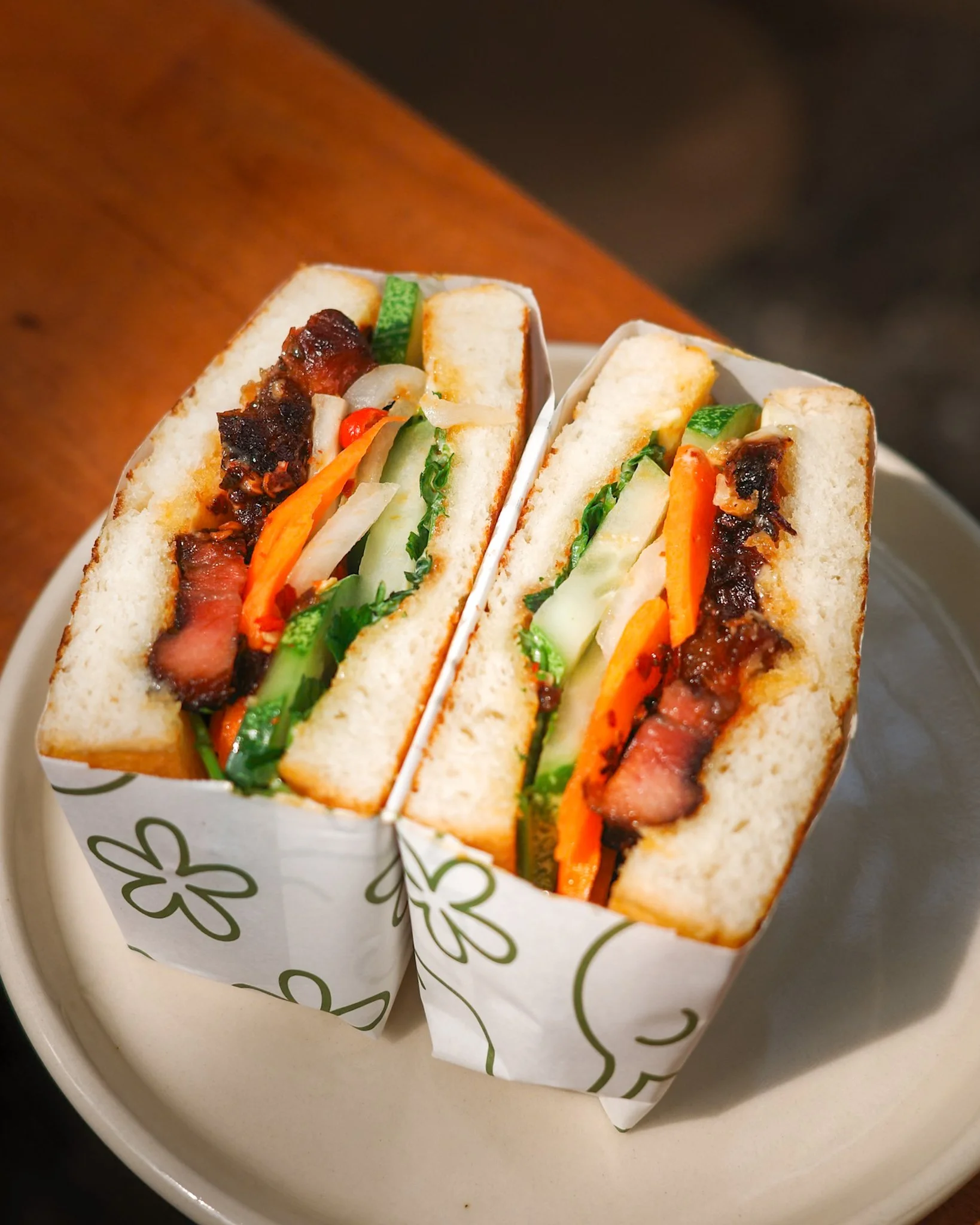 Close-up of two Vietnamese-style banh mi sandwiches with grilled pork, vegetables, and herbs in paper wrappers on a white plate.
