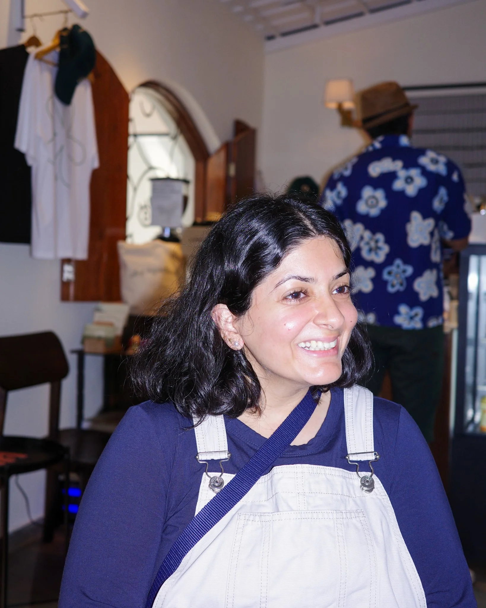 A woman with shoulder-length black hair smiling, wearing a navy blue top and beige overalls, in a casual indoor setting.