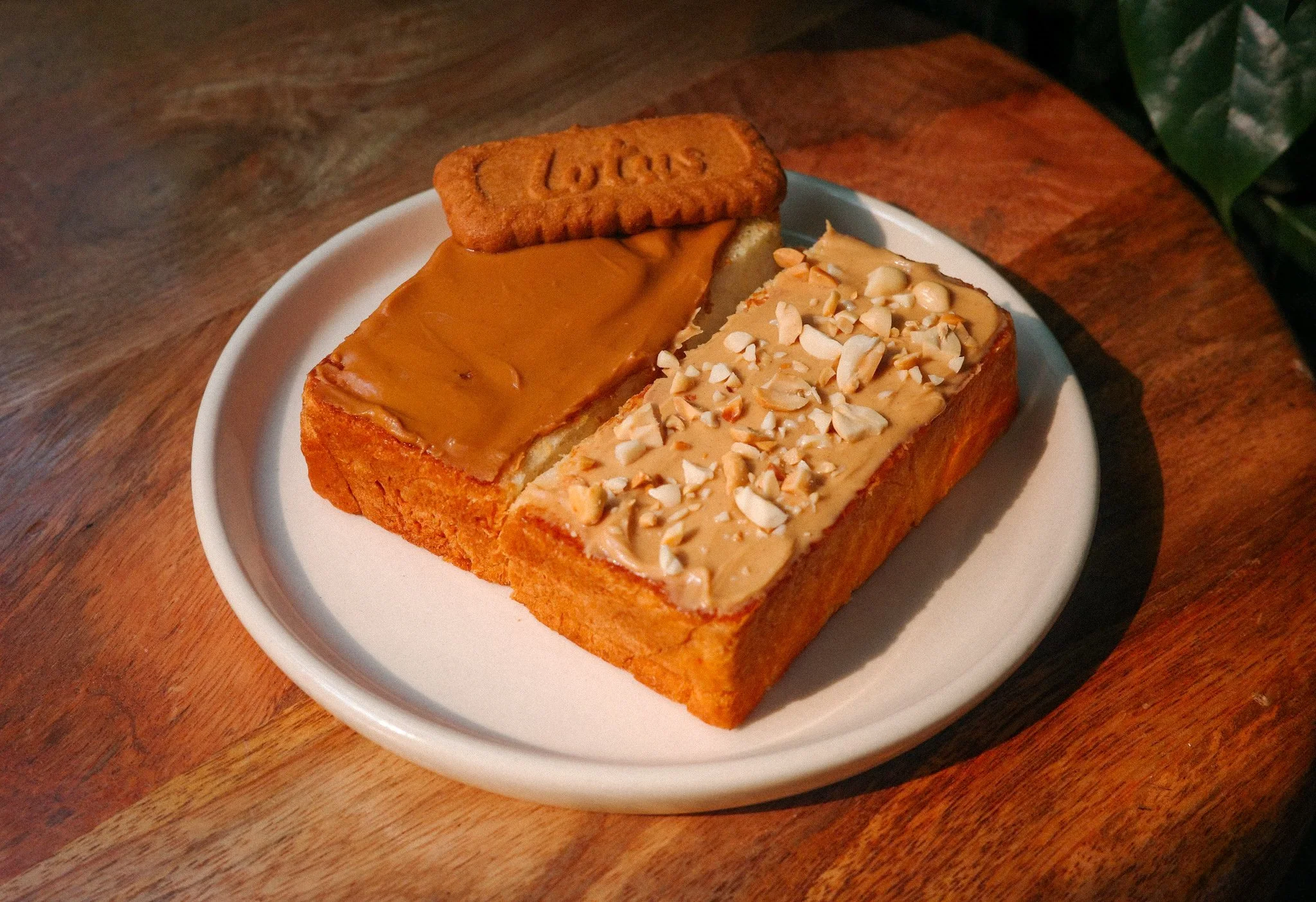 Two slices of bread topped with peanut butter, one topped with caramel and the other with chopped nuts, and a biscuit on a white plate on a wooden table.
