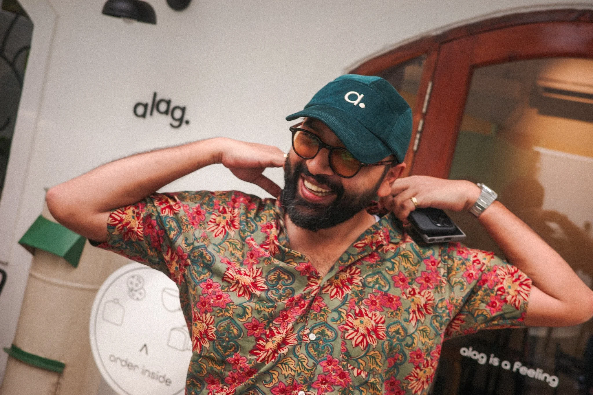 A man with a beard and glasses wearing a dark blue cap with a white lowercase 'a' and a colorful floral shirt, smiling and adjusting his cap while holding a phone to his ear.