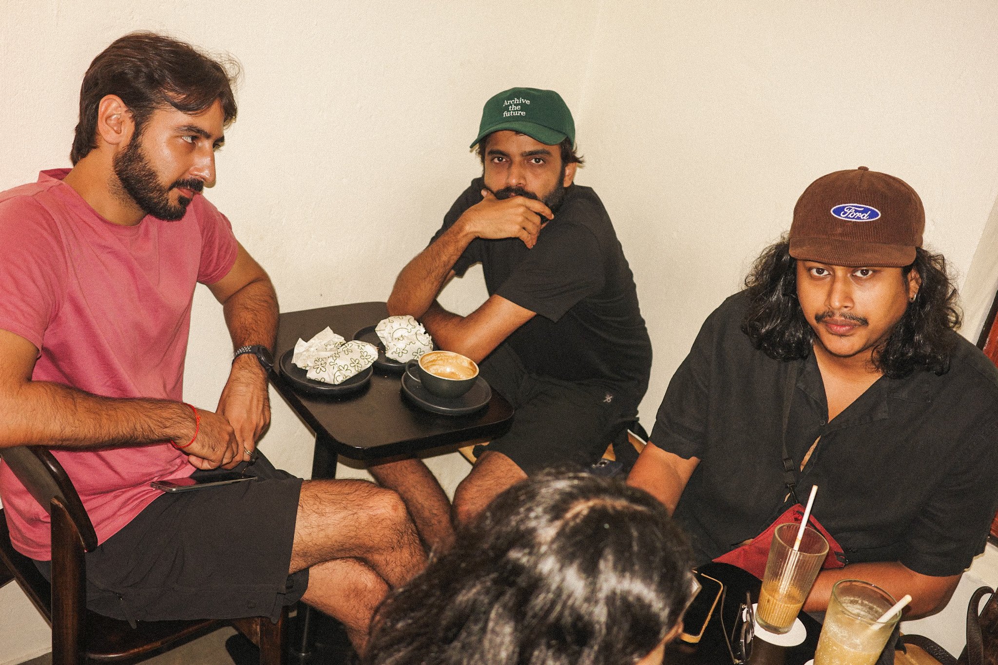 Three young men sitting around a small table in a café or restaurant, with cups of coffee and drinks, and a woman with black hair partially visible in the foreground.