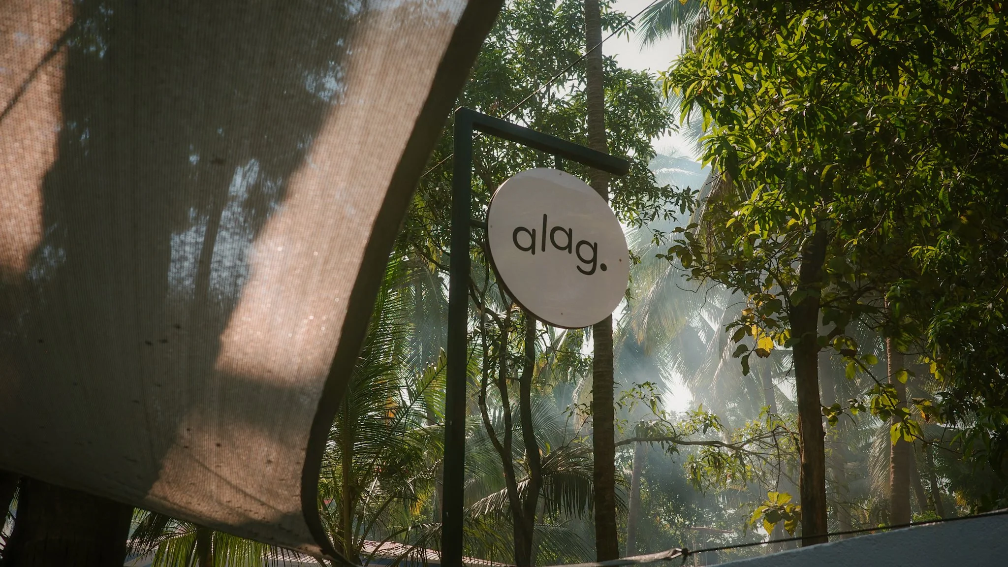 Outdoor scene with a circular white sign that says 'alag.' in black lowercase letters, framed by green tropical trees and sunlight filtering through the leaves.