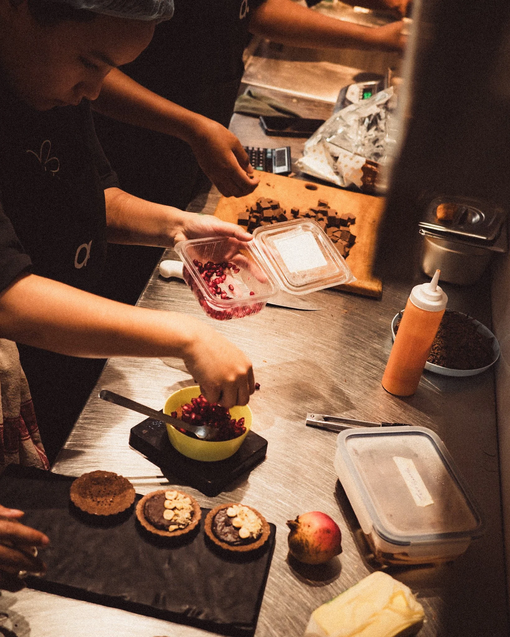 People preparing and decorating desserts in a kitchen, with plates of cookies, a bowl of pomegranate seeds, an apple, and a block of butter on the counter.