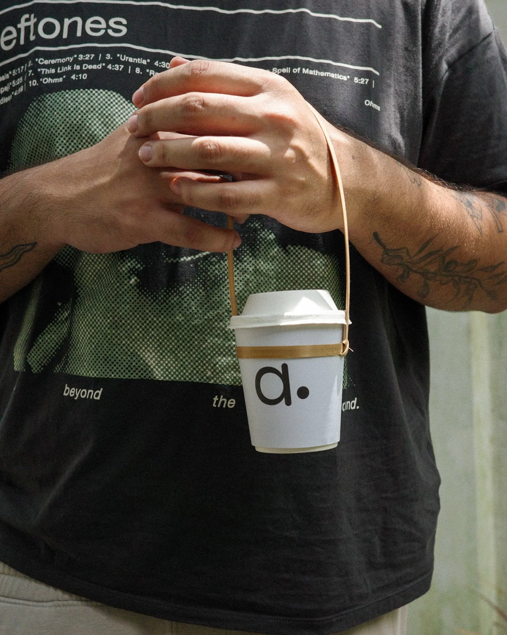 Person with tattoos holding a coffee cup with a string handle, hanging from their hands.