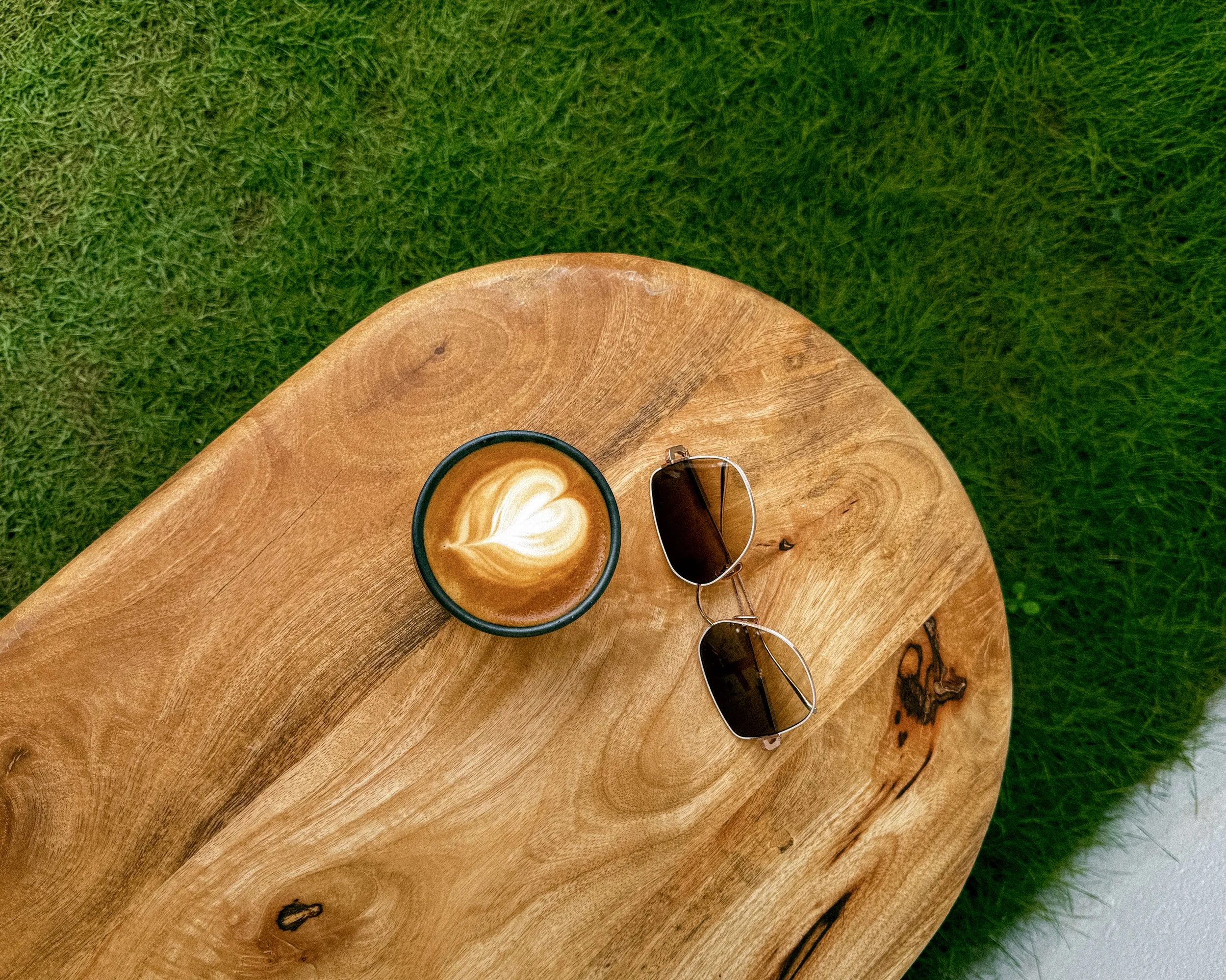 A wooden table with a cup of latte art and sunglasses on top, set on green grass.