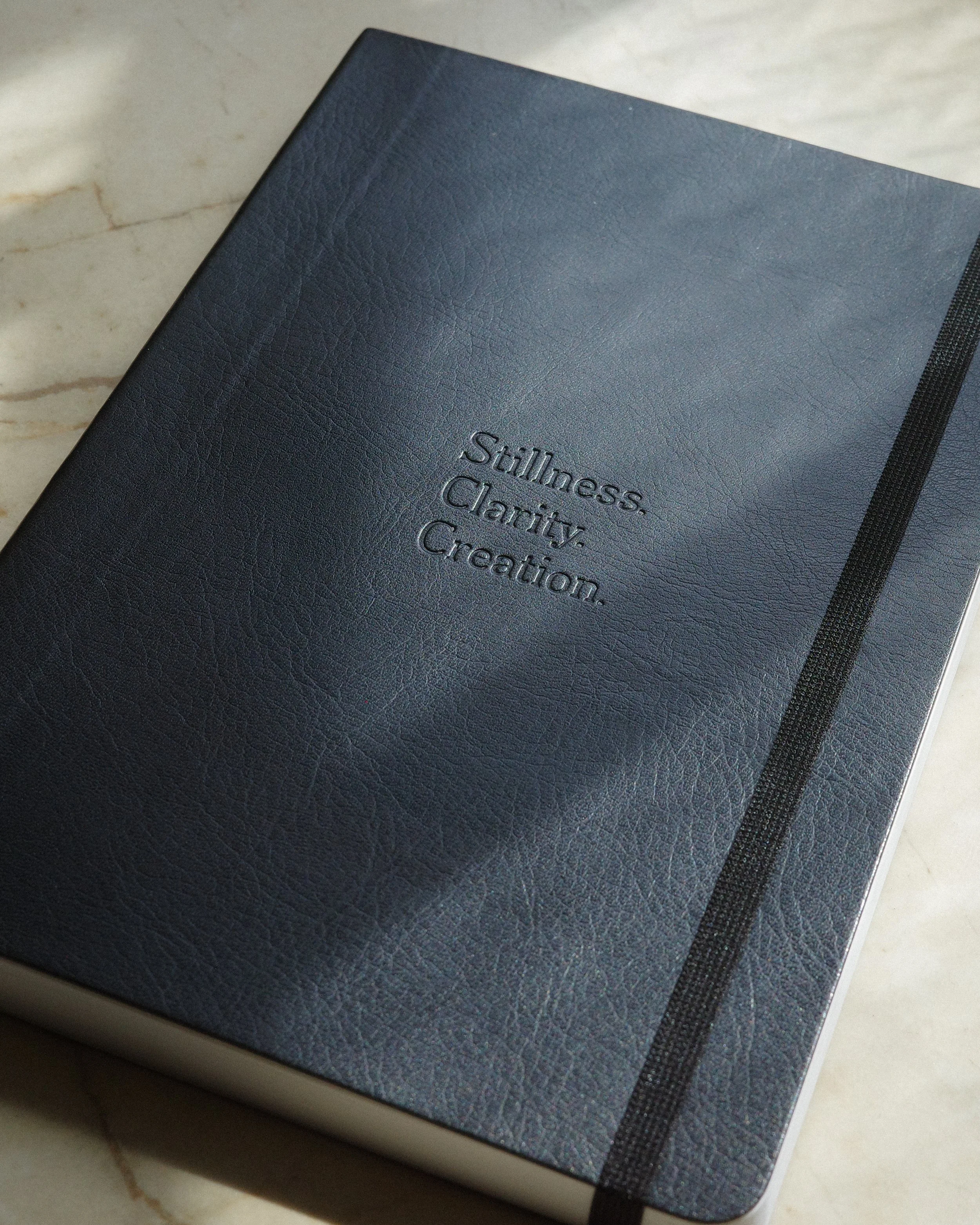 A navy blue leather-bound notebook with the words "Stillness. Clarity. Creation." embossed on the cover, placed on a light-colored marble surface.