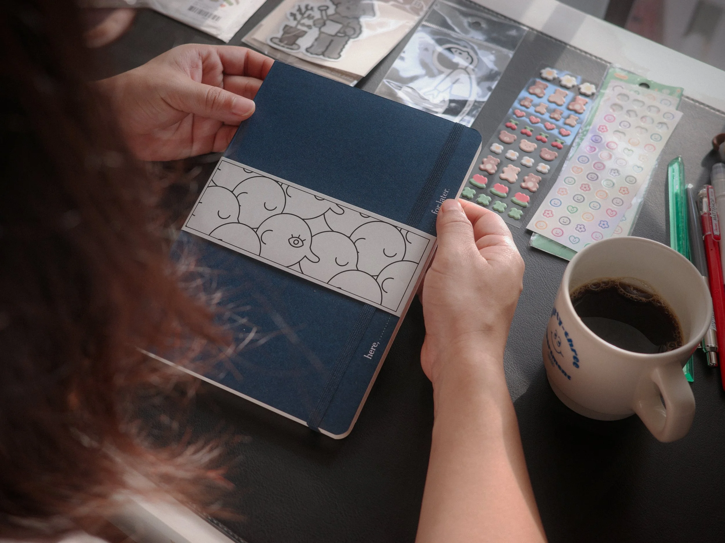 A person sitting at a table with a dark surface, holding a closed navy blue notebook with a sticker of cartoon smiling cherries on it. Next to the person are stickers, pens, and a mug of coffee.